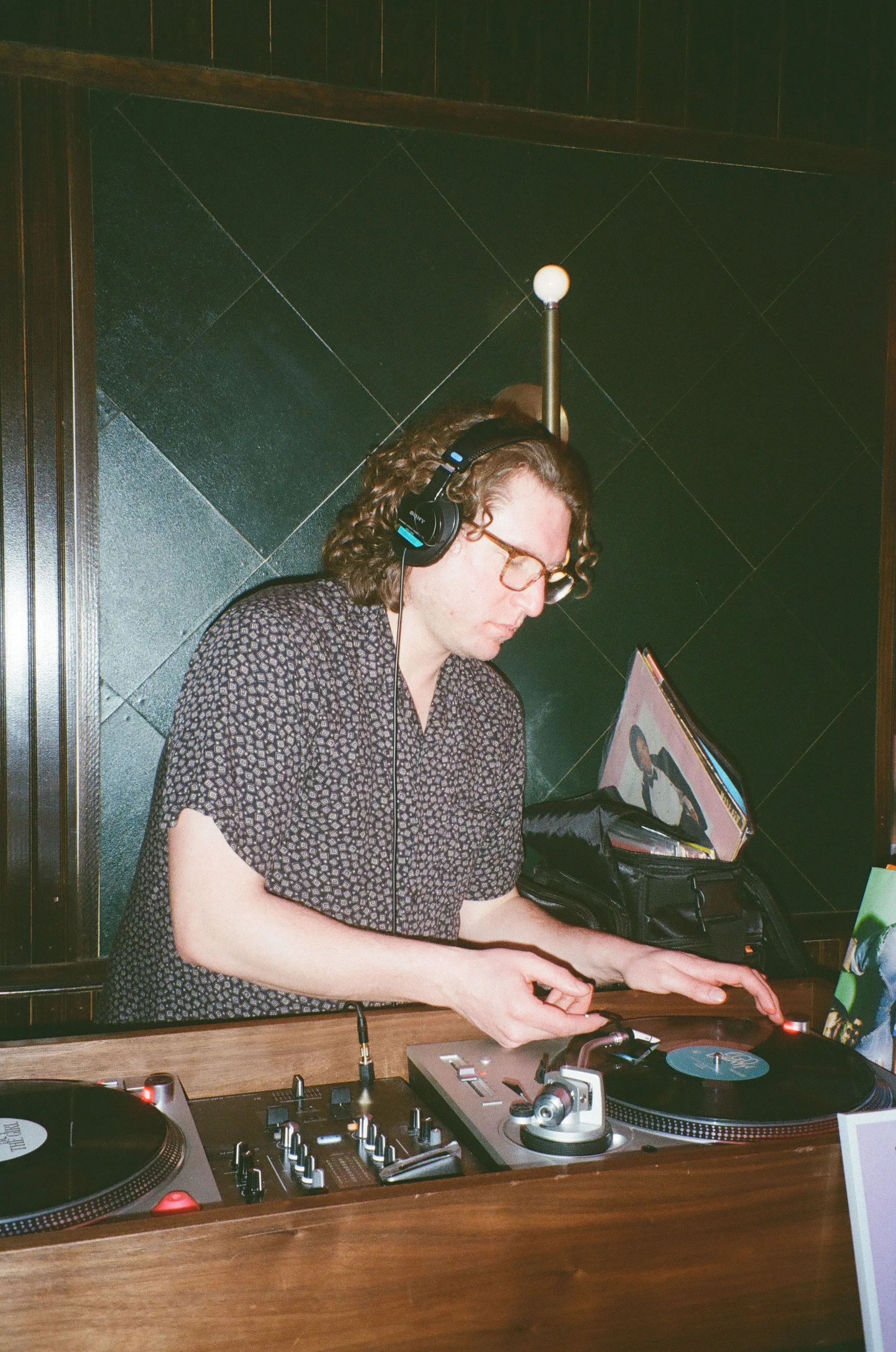A woman with curly hair and glasses wearing headphones, mixing music on DJ equipment at a wooden table in a dimly lit room.