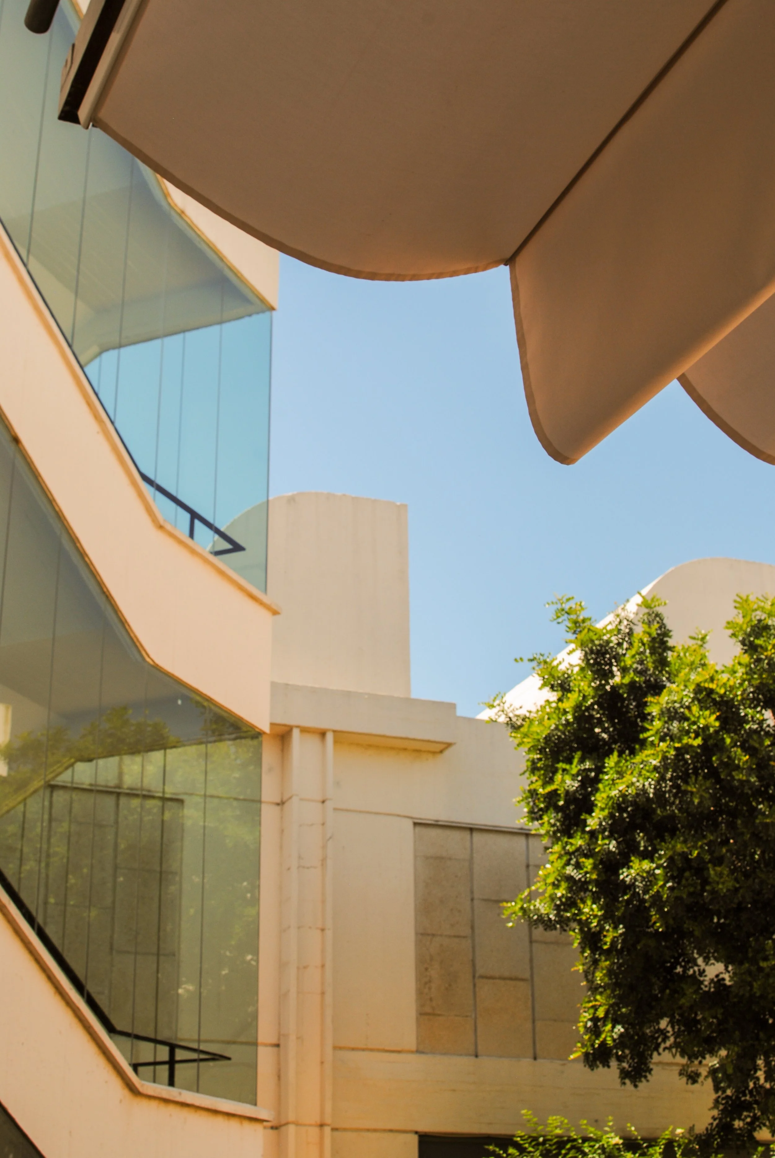 Modern building with large glass windows, beige walls, and curved architectural features, partially covered by large beige umbrellas or canopies, with trees and clear blue sky in the background.