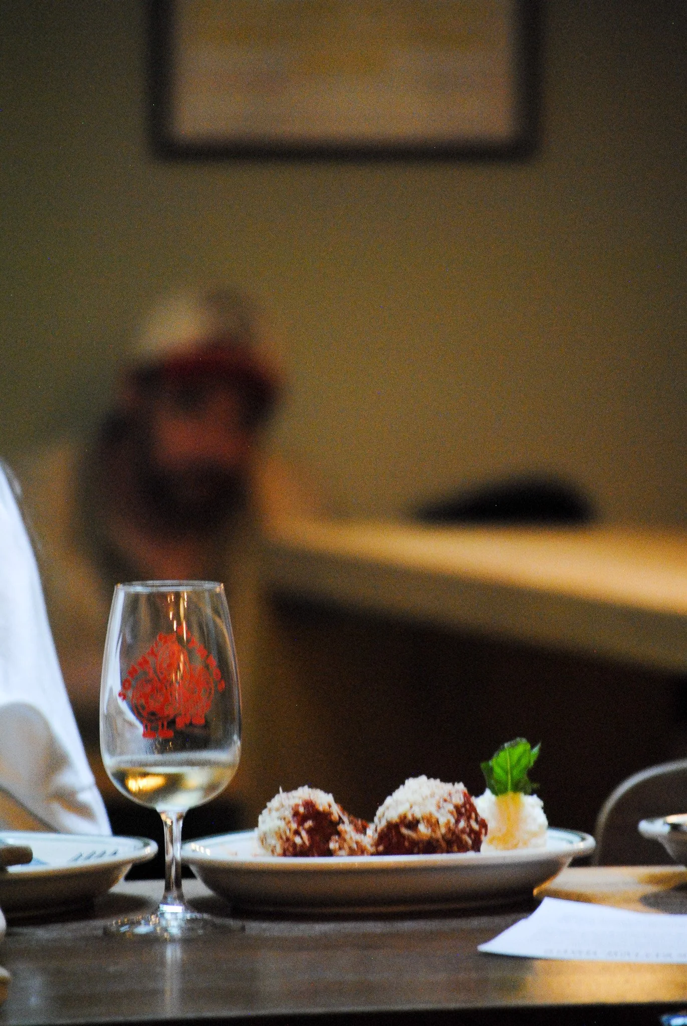 A table with a glass of white wine, a plate of breaded and fried food with sauces, and some green garnishes in a restaurant setting. In the background, there is a blurry figure and a picture frame on the wall.