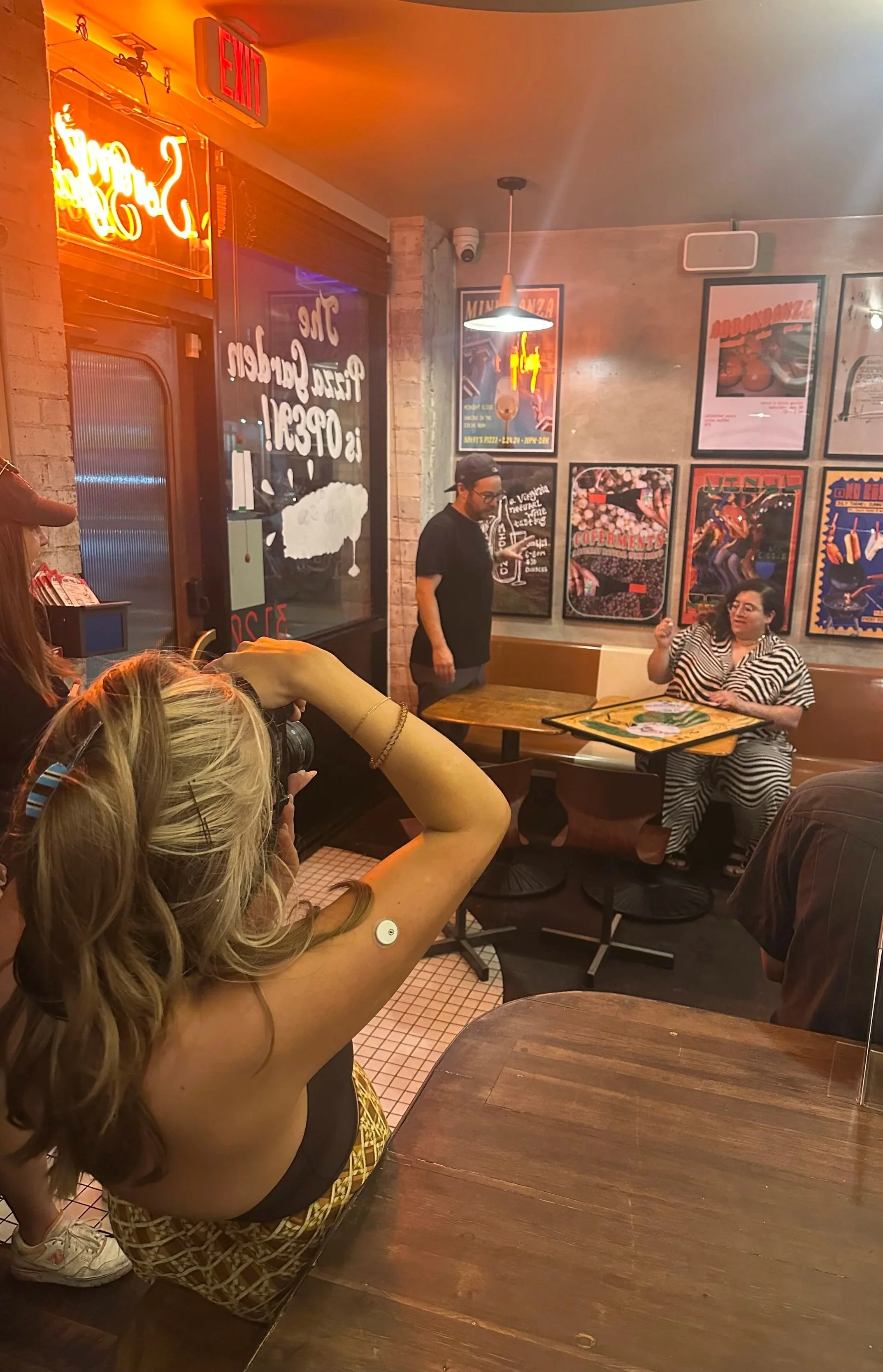 Interior of a restaurant with brick walls, posters on the wall, and a group of people. A woman with blonde hair, wearing a black top with spaghetti straps and patterned shorts, is in the foreground taking a photo or video. A man in a black shirt and glasses is standing near a table, and another woman with dark hair and striped dress is seated at a table. Neon signs and the restaurant's window with writing are visible.