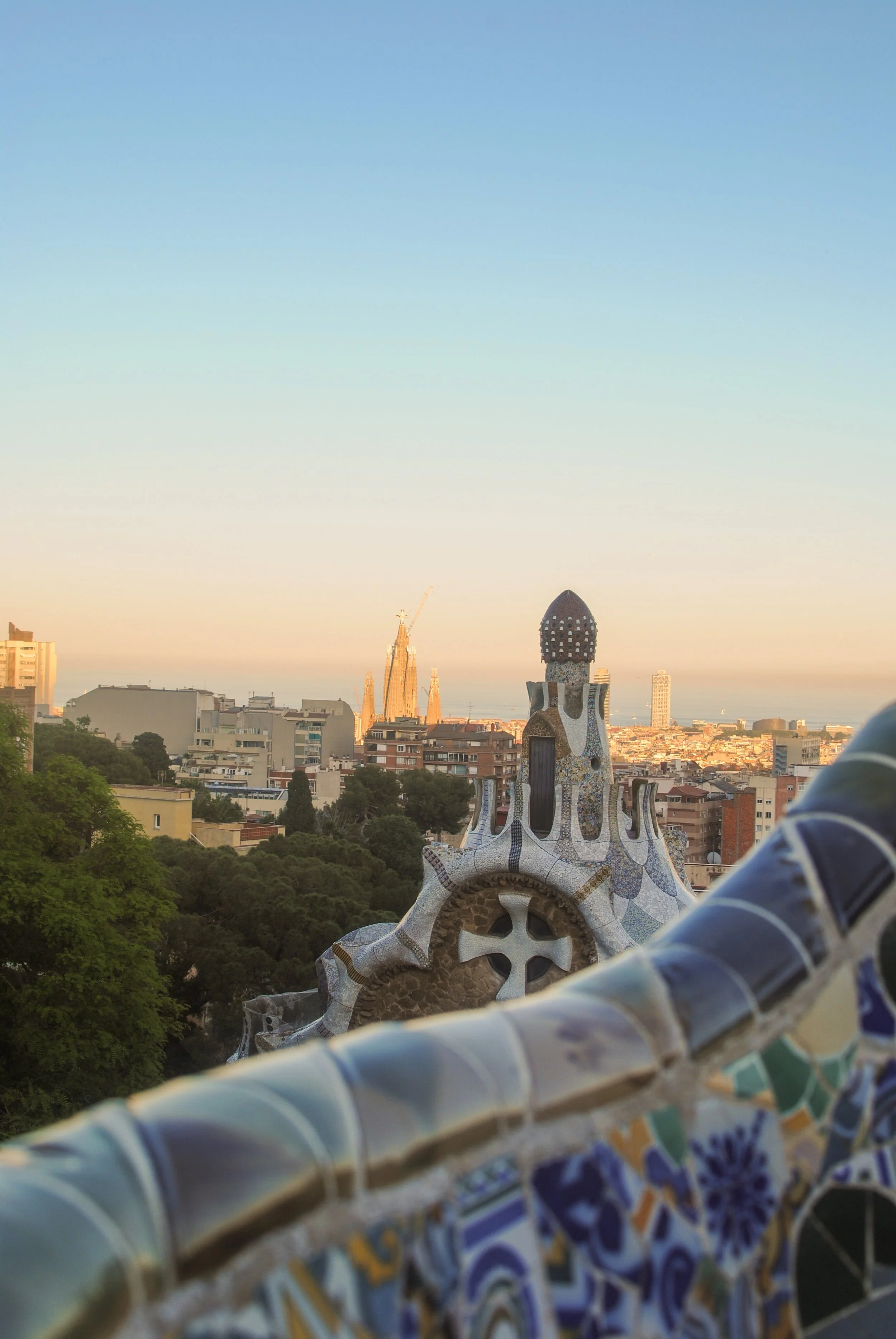 View of Barcelona city skyline during sunset, with a mosaic-covered rooftop structure in the foreground and the Sagrada Família basilica in the distance.