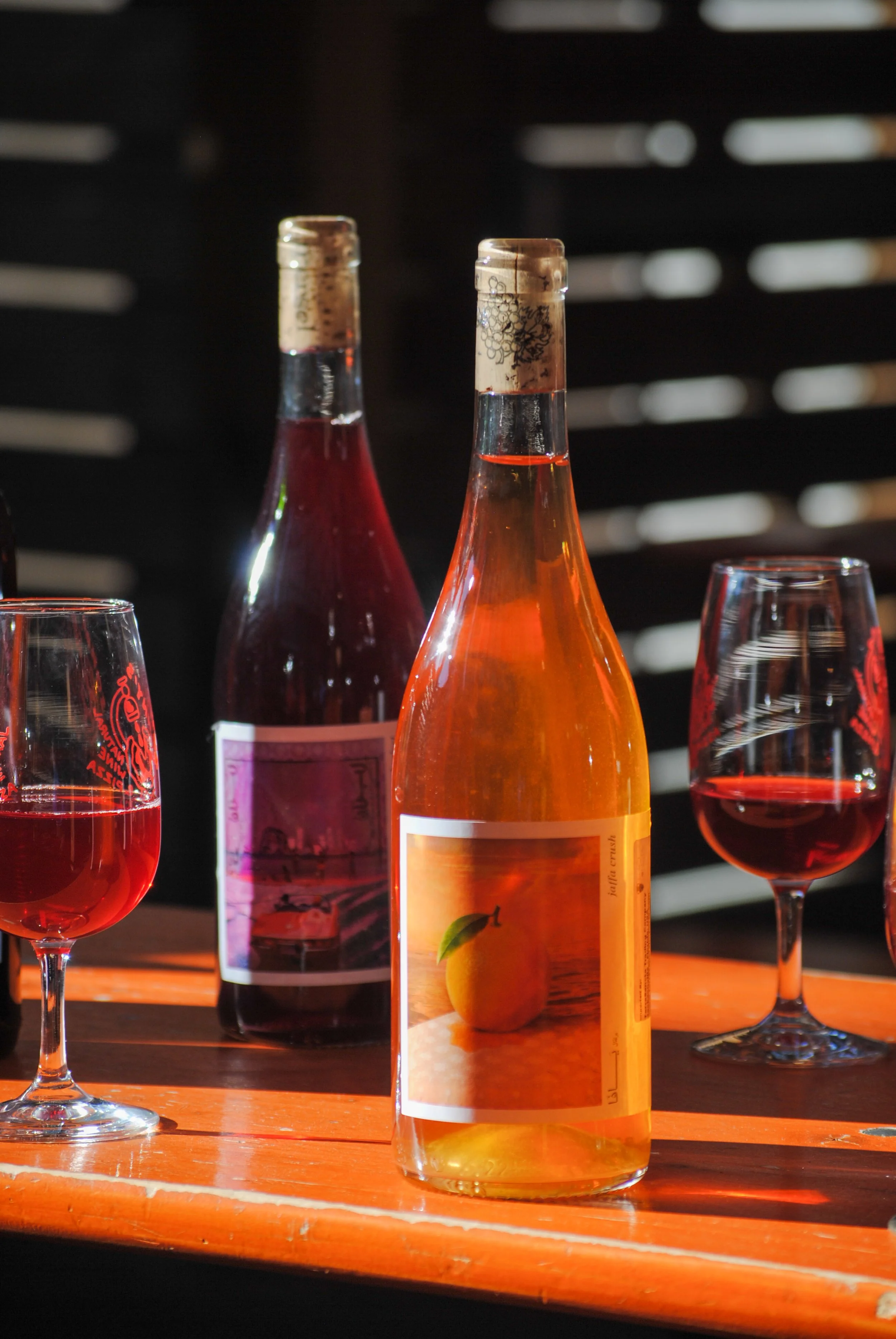 Two bottles of rosé wine and two wine glasses filled with rosé on a wooden table, with a black slatted background.
