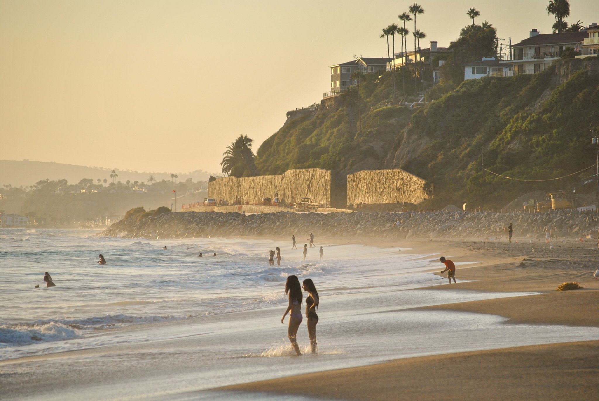 Beach scene during sunset with people swimming and walking along the shoreline, green hillside with residential houses and tall palm trees in the background.