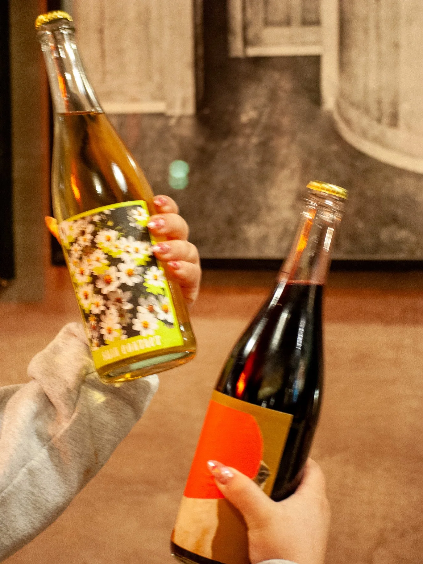 Two hands holding a bottle of wine and a bottle of sparkling cider, with a kitchen background.