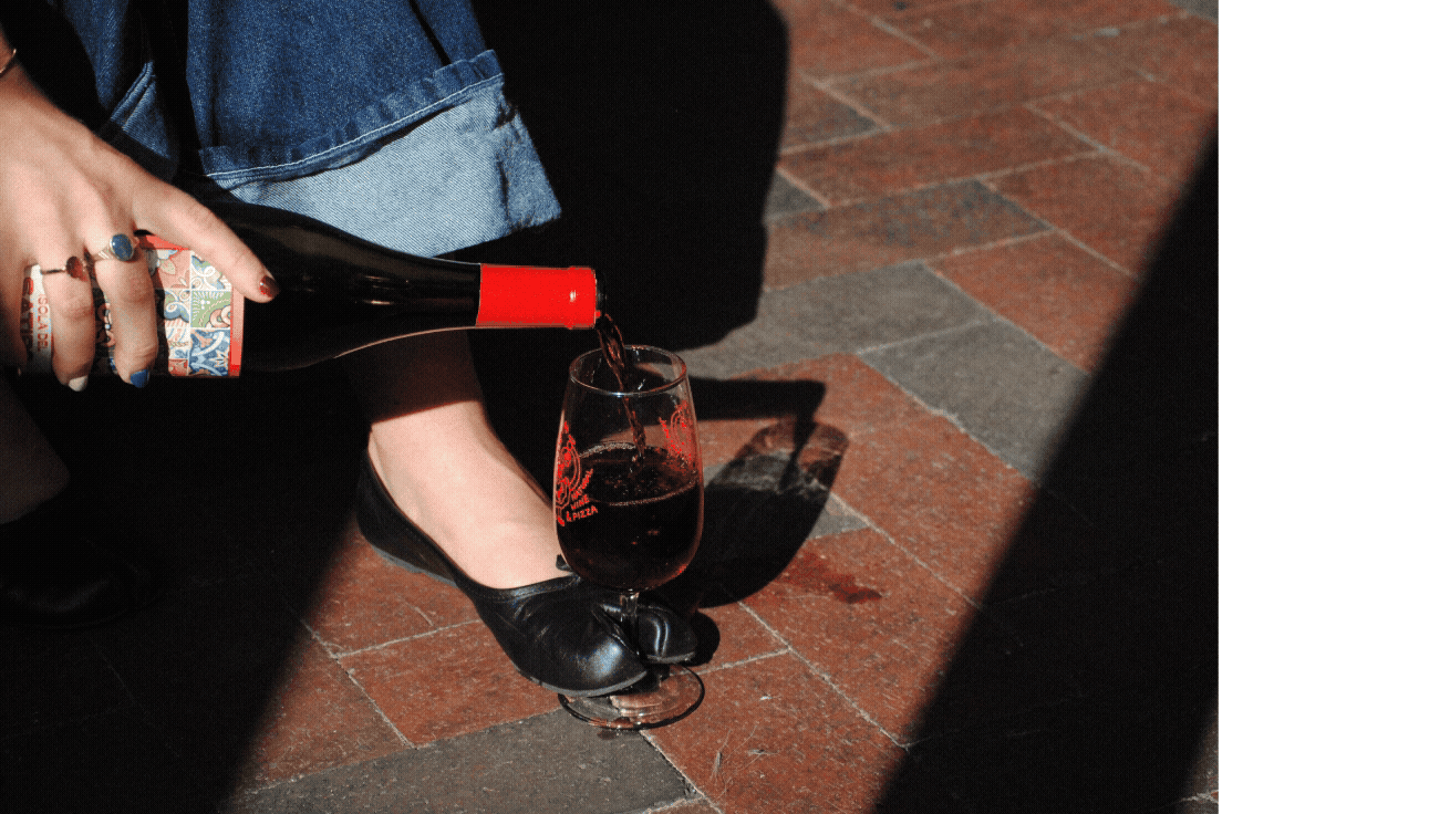 Person pouring red wine into a glass while sitting on a brick floor, wearing black shoes and blue jeans.