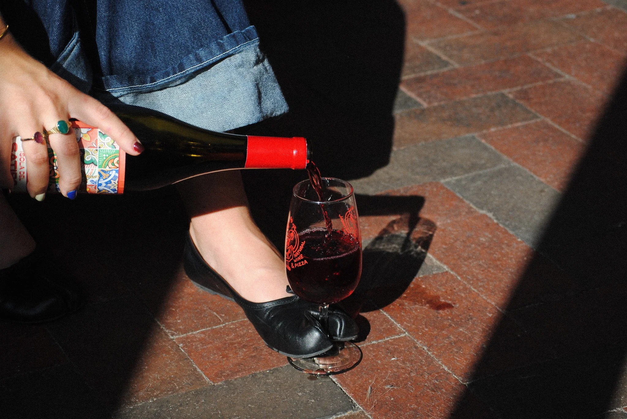 Person pouring red wine from a bottle into a wine glass on a brick floor, wearing rings and a denim shirt.