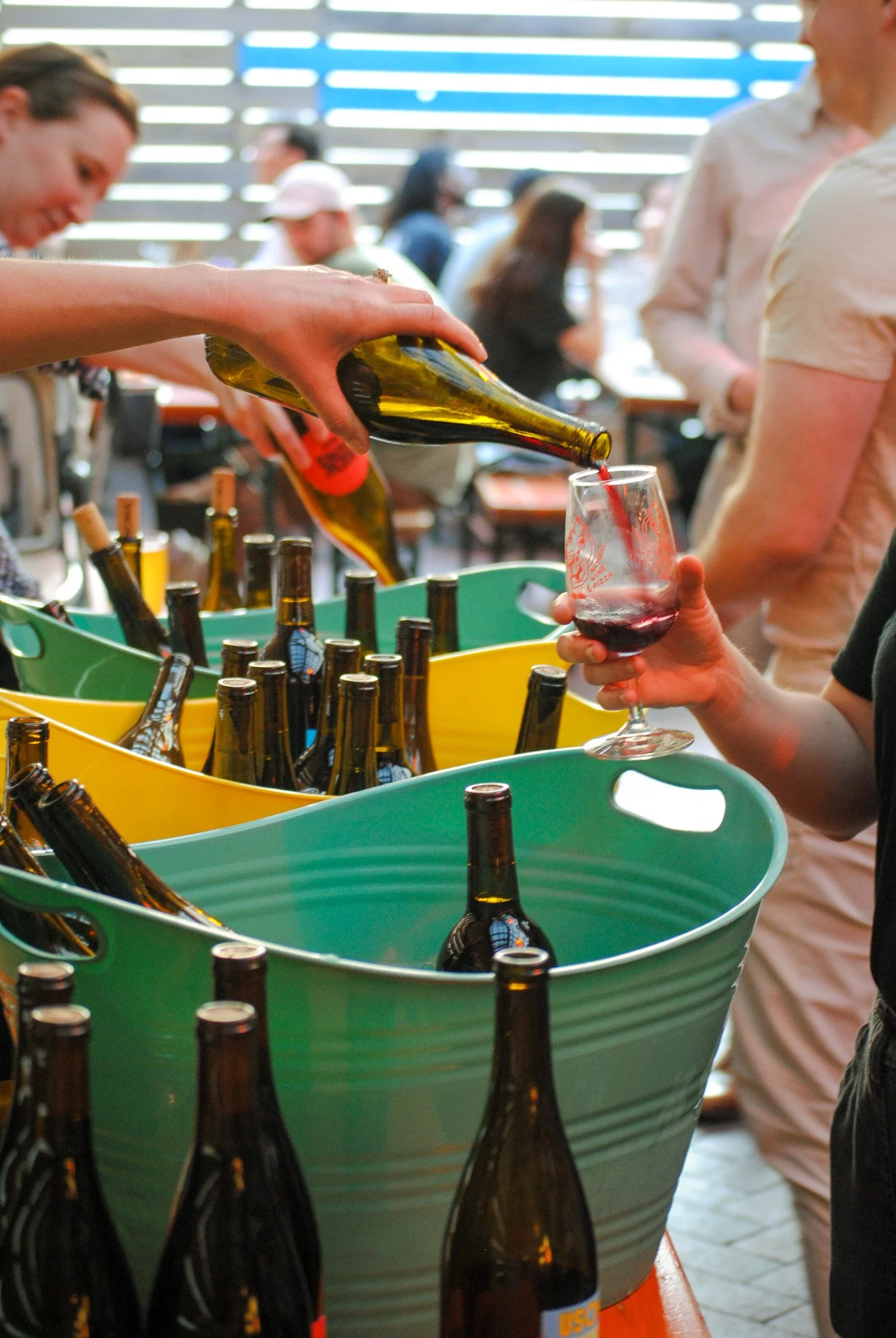 Person pouring red wine from a green bottle into a wine glass at a social gathering with people in the background.
