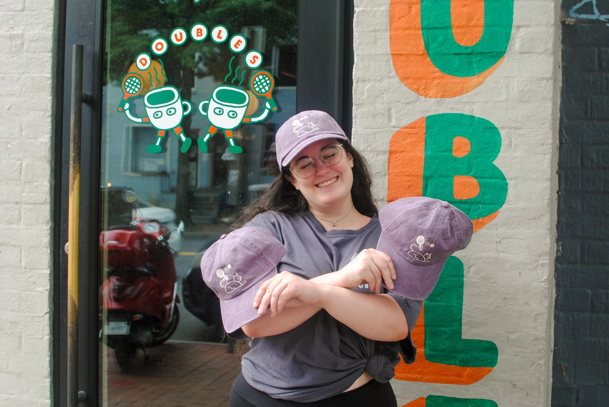 A person with glasses and a purple cap smiling with eyes closed, standing in front of a door with a colorful sign that says "Doughnuts" and has two cartoon coffee cups holding steaming doughnuts.