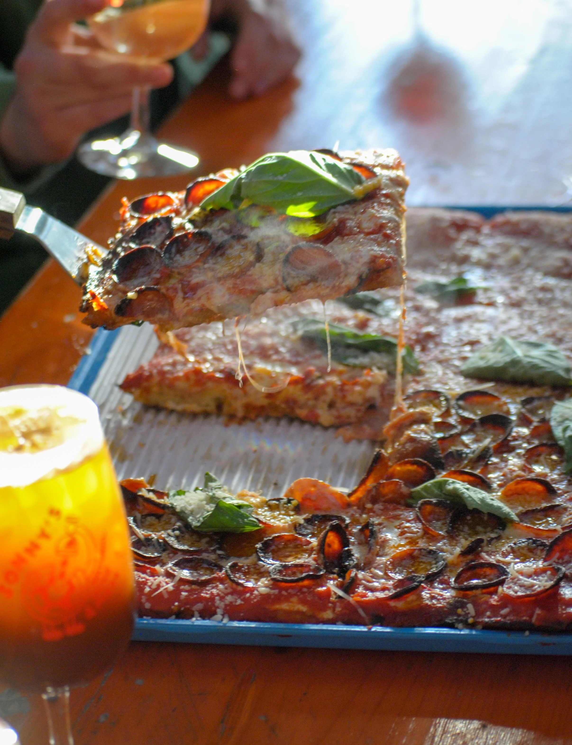 Close-up of a slice of pizza with pepperoni and basil leaves, with a person's hand holding a wine glass in the background.