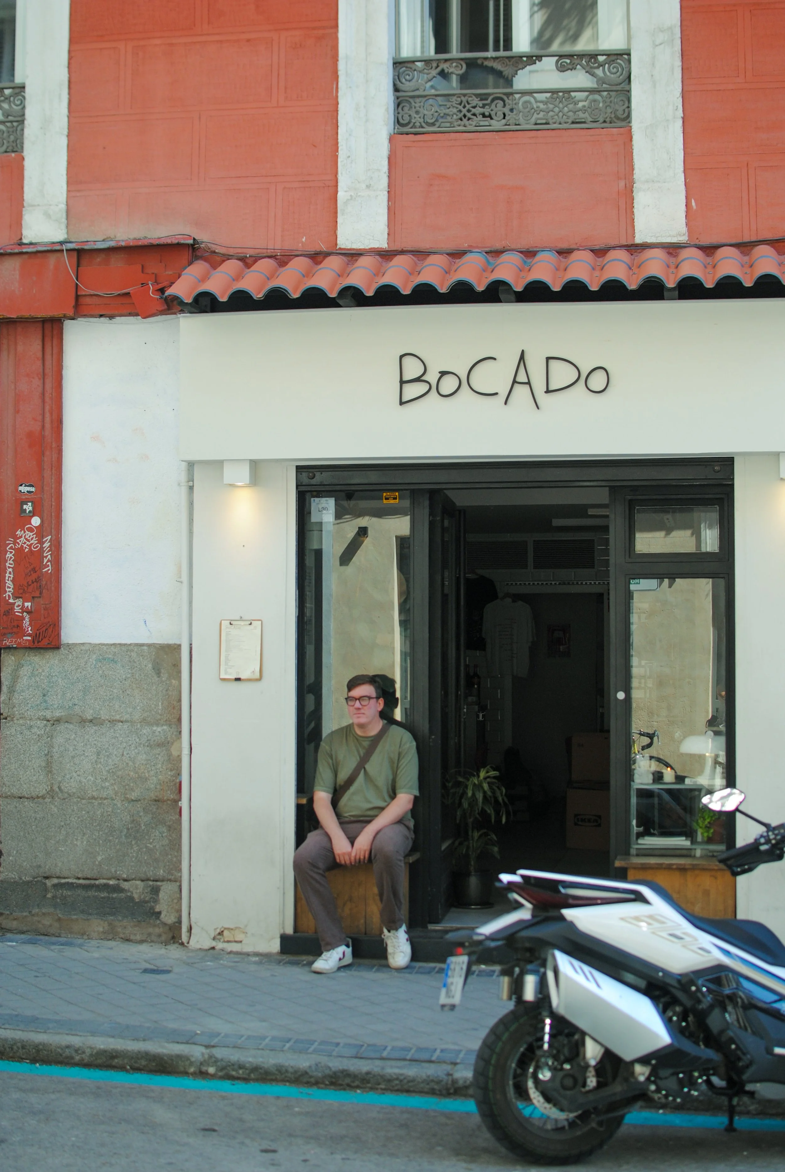A man sitting outside a cafe or shop, wearing glasses, a green shirt, brown pants, and white sneakers. Behind him, the building has a white storefront with the sign 'BoCADO' and a motorcycle parked on the street in front.