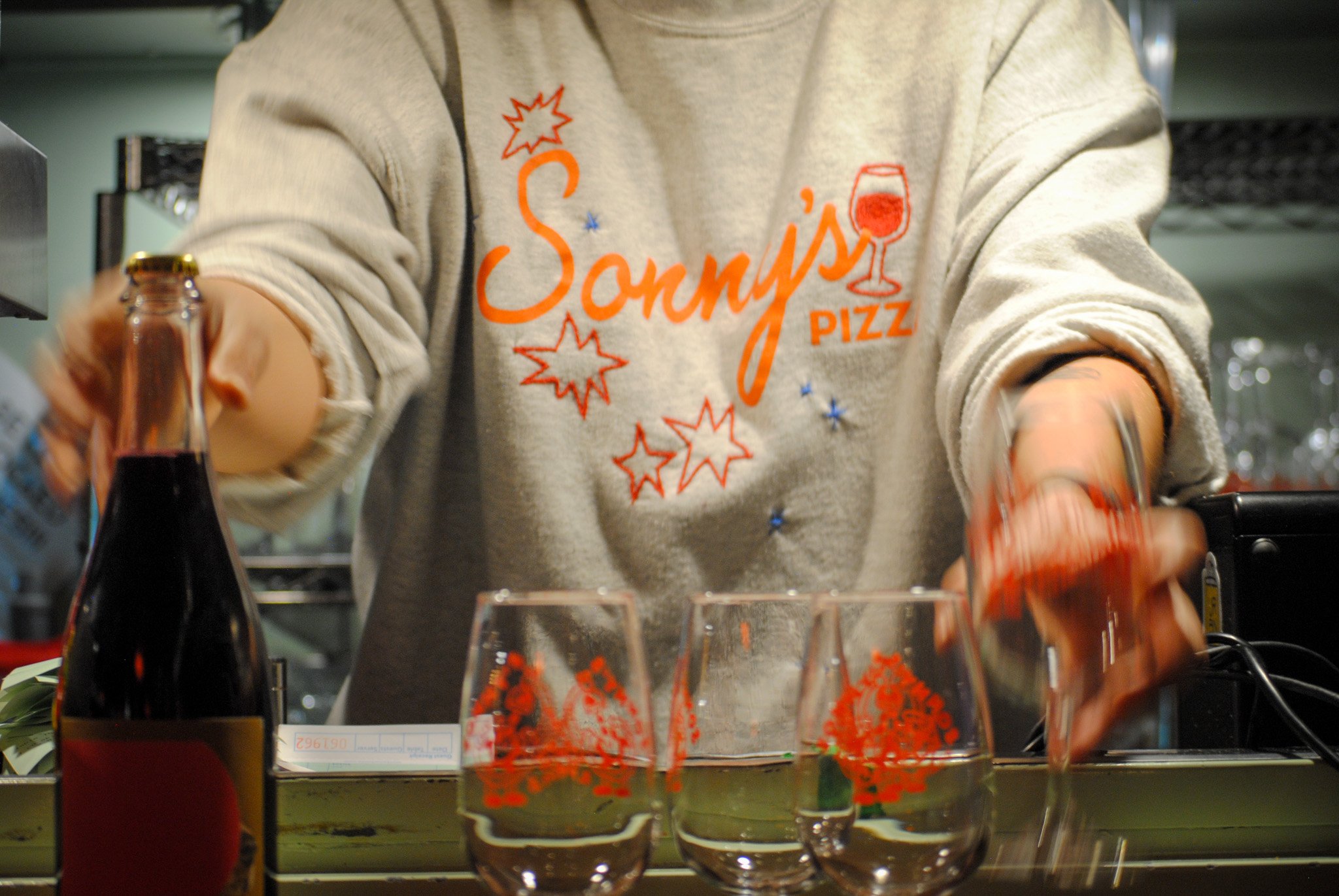 A person in a gray sweatshirt with orange and red graphics serving drinks at a bar or restaurant. The sweatshirt has a glass of wine and starburst designs. There are several glasses with red wine and a soda bottle on the table.