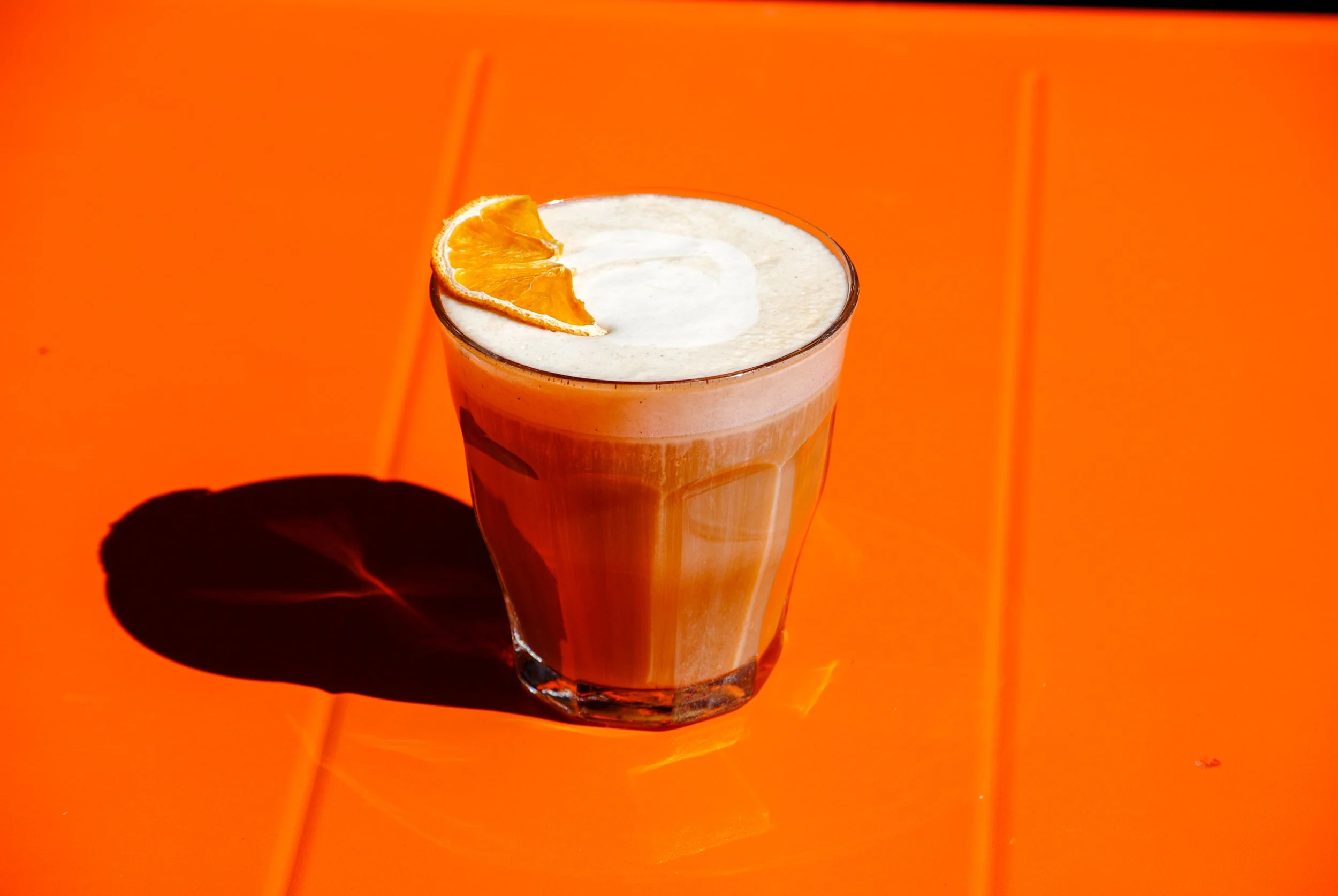 A tall glass of orange juice garnished with an orange slice on the rim, casting a shadow on an orange background.