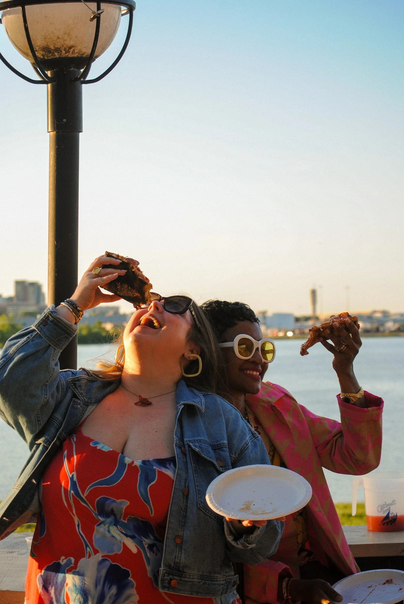 Three women enjoying pizza outdoors near a body of water, with a city skyline in the background, during sunset.
