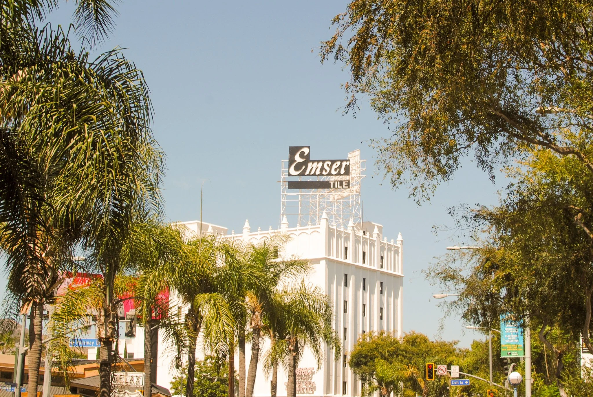 Street scene with tall palm trees and a large white building in the background, featuring a sign that reads 'Emser Tile' open sky and trees around.
