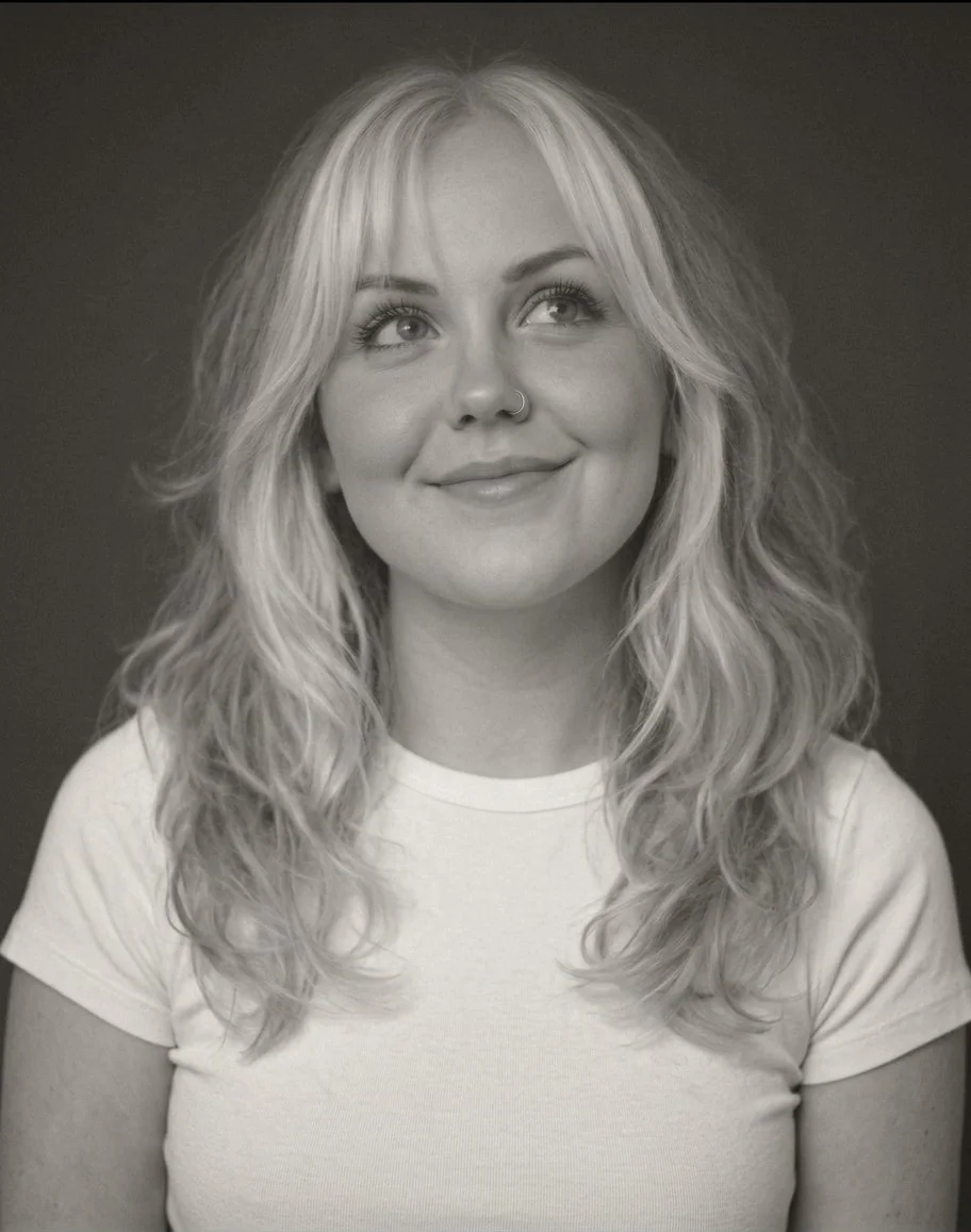 Black and white portrait of a young woman with wavy blonde hair, smiling while looking up and to the side, wearing a plain light-colored t-shirt.