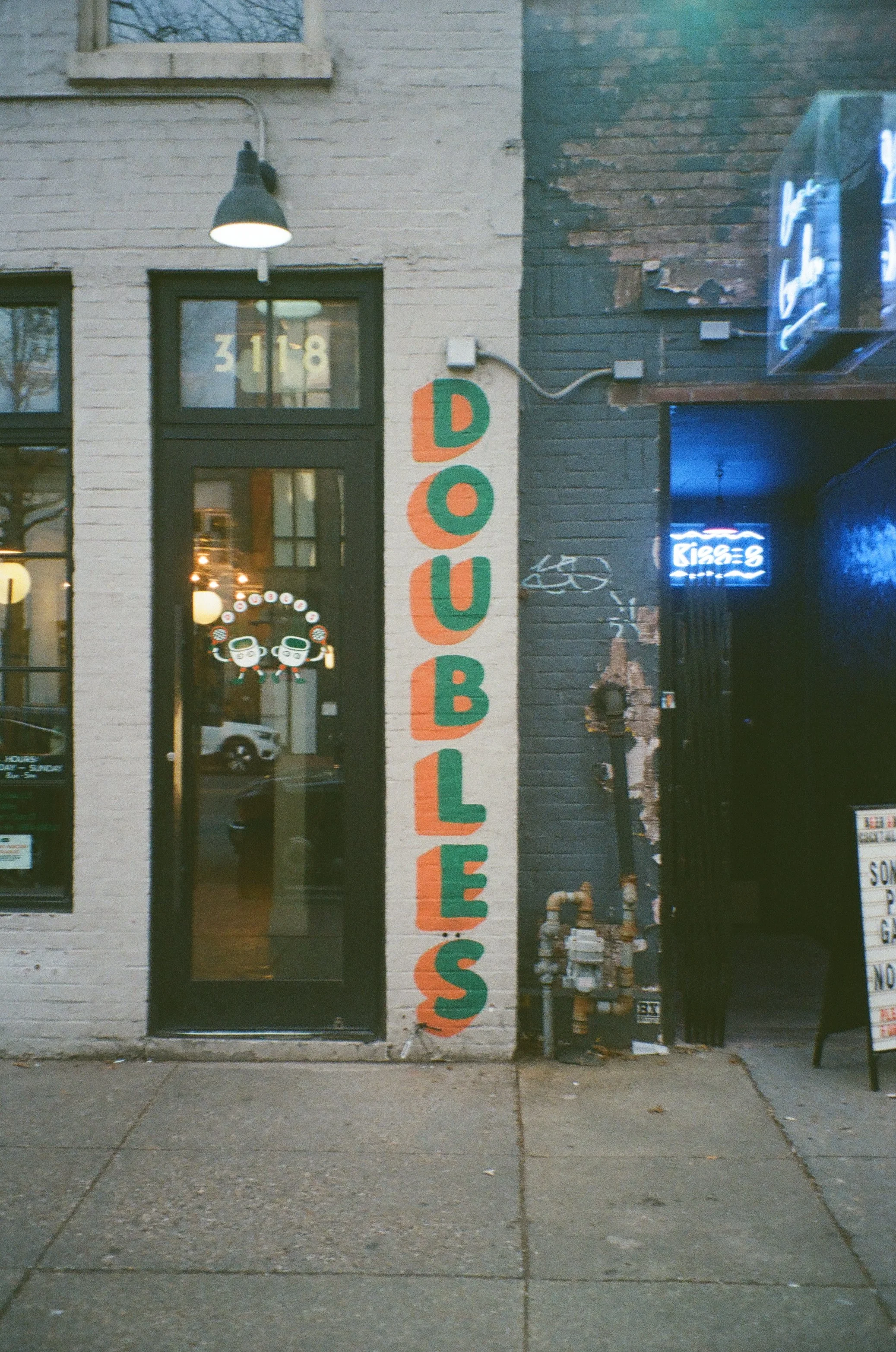 A storefront with a sign reading 'Dough' painted in colorful letters on a vertical white board. The door has a sticker of two happy cartoon sushi rolling characters. To the right, part of a neon sign is visible, and the building features a combinatio