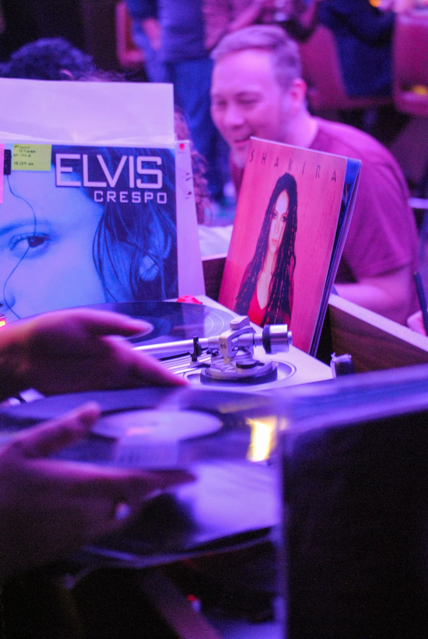 A person is DJing with a hand on turntable, with vinyl records and music albums, including Elvis Crespo and Shakira, visible in the background, illuminated by purple and pink lighting.