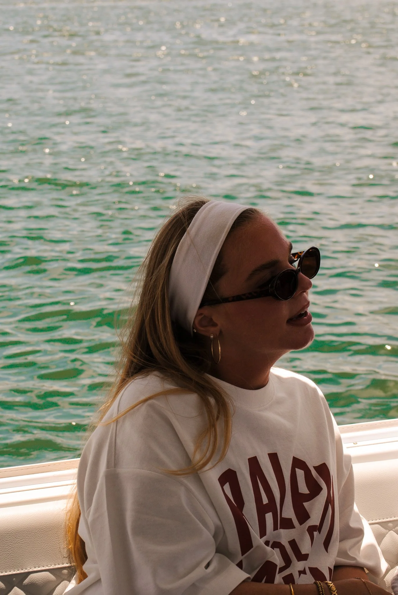 A woman with long hair, wearing sunglasses, a headband, earrings, and a white sweatshirt, sitting on a boat with water in the background.