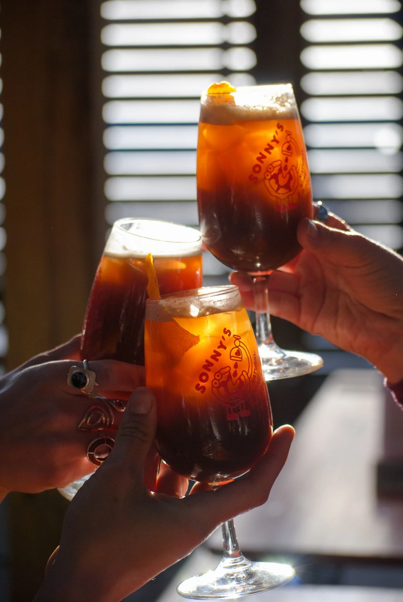 Multiple hands holding glasses of dark amber beer with foam on top, against a window with blinds, celebrating with cheers.