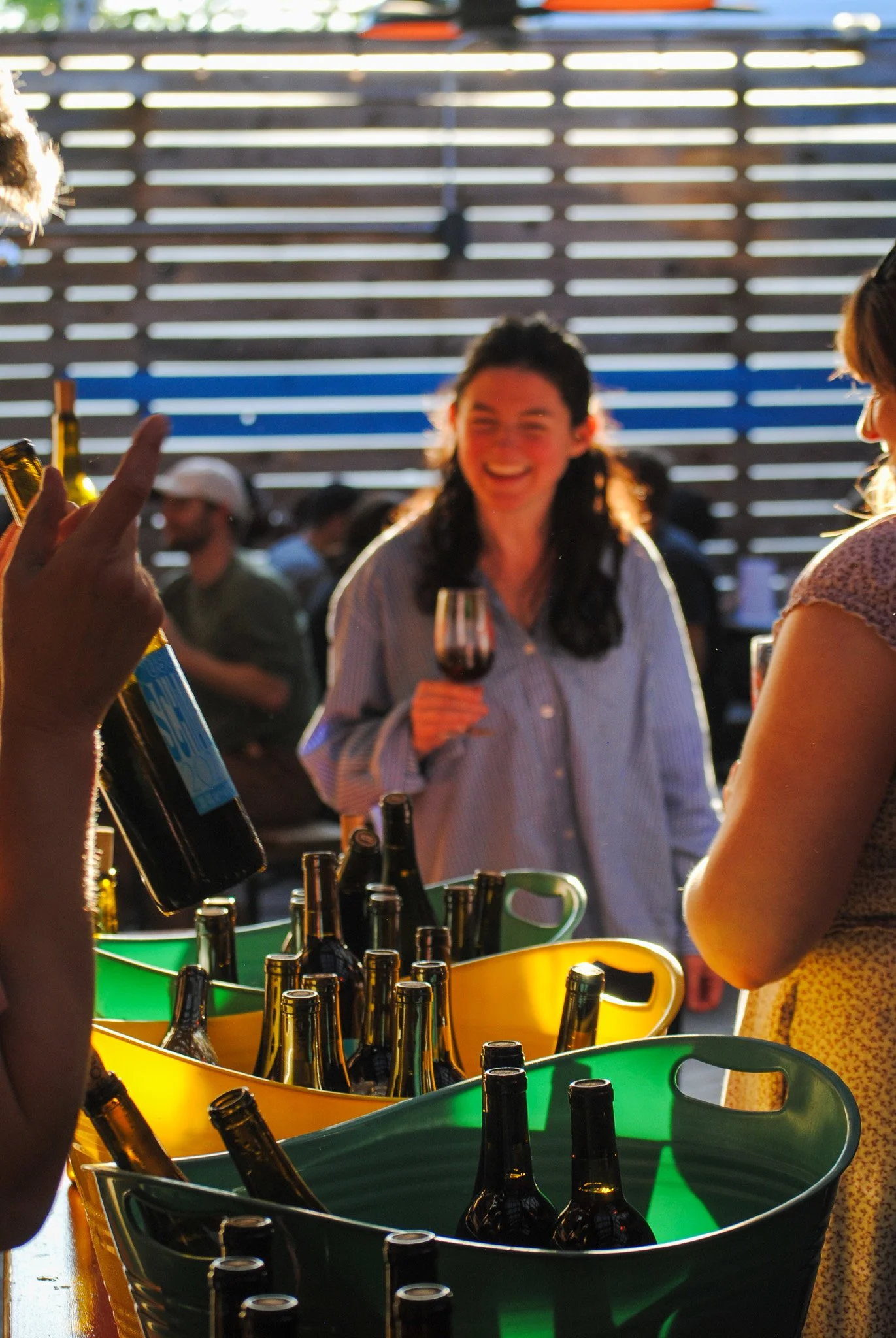 People socializing outdoors, with beer bottles in colorful buckets in the foreground, and a woman smiling holding a wine glass in the background.