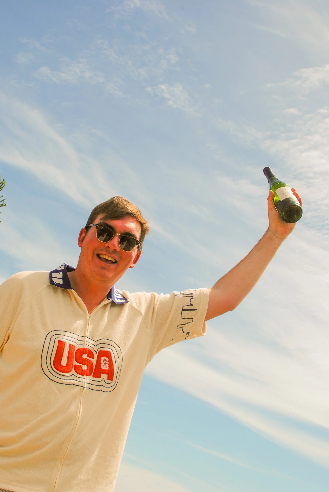 A man with short brown hair and sunglasses smiling while holding a green bottle in his right hand, wearing a light yellow USA racing jacket, against a blue sky with some scattered clouds.