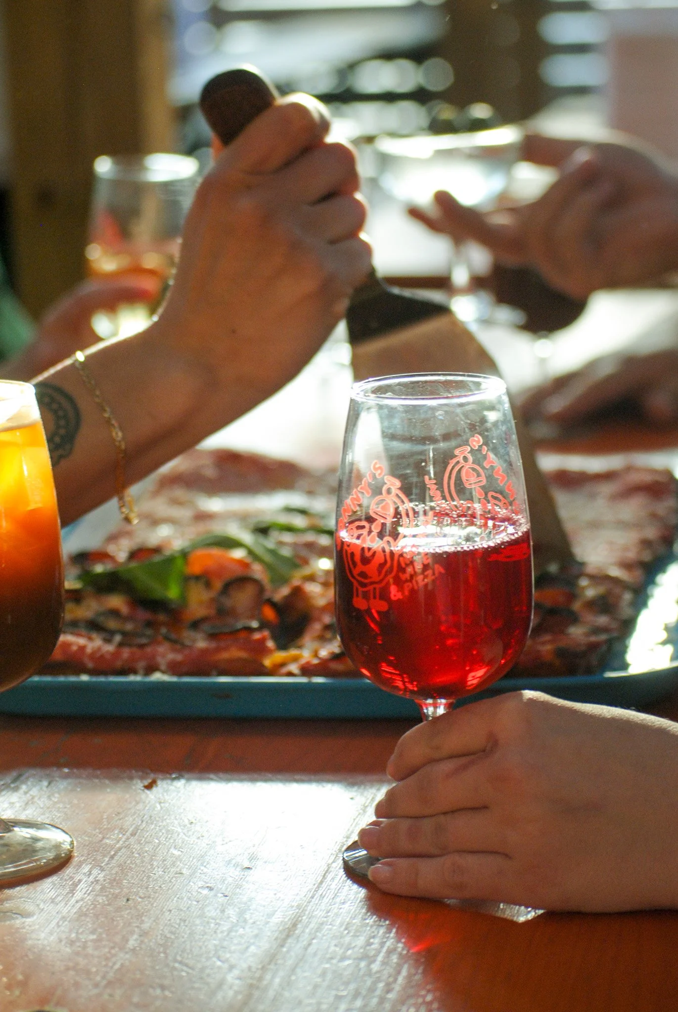 Close-up of a person holding a glass of red wine, with pizza and food on the table in the background.