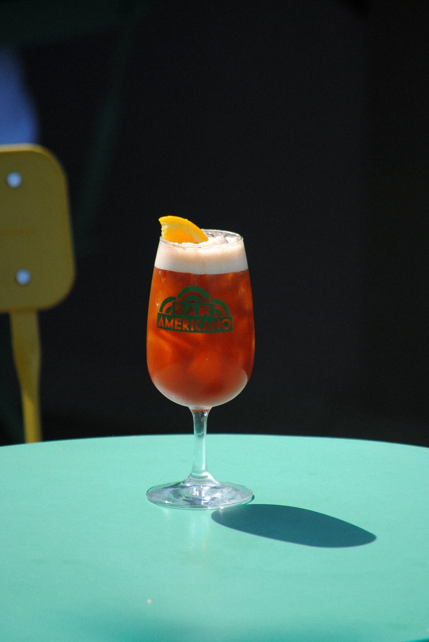A cocktail glass with a red drink and a frothy top, garnished with an orange wedge, placed on a light green table against a dark background.