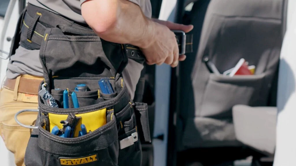 A person wearing a tool belt with various blue-handled tools, holding a smartphone, standing inside or near an airplane.