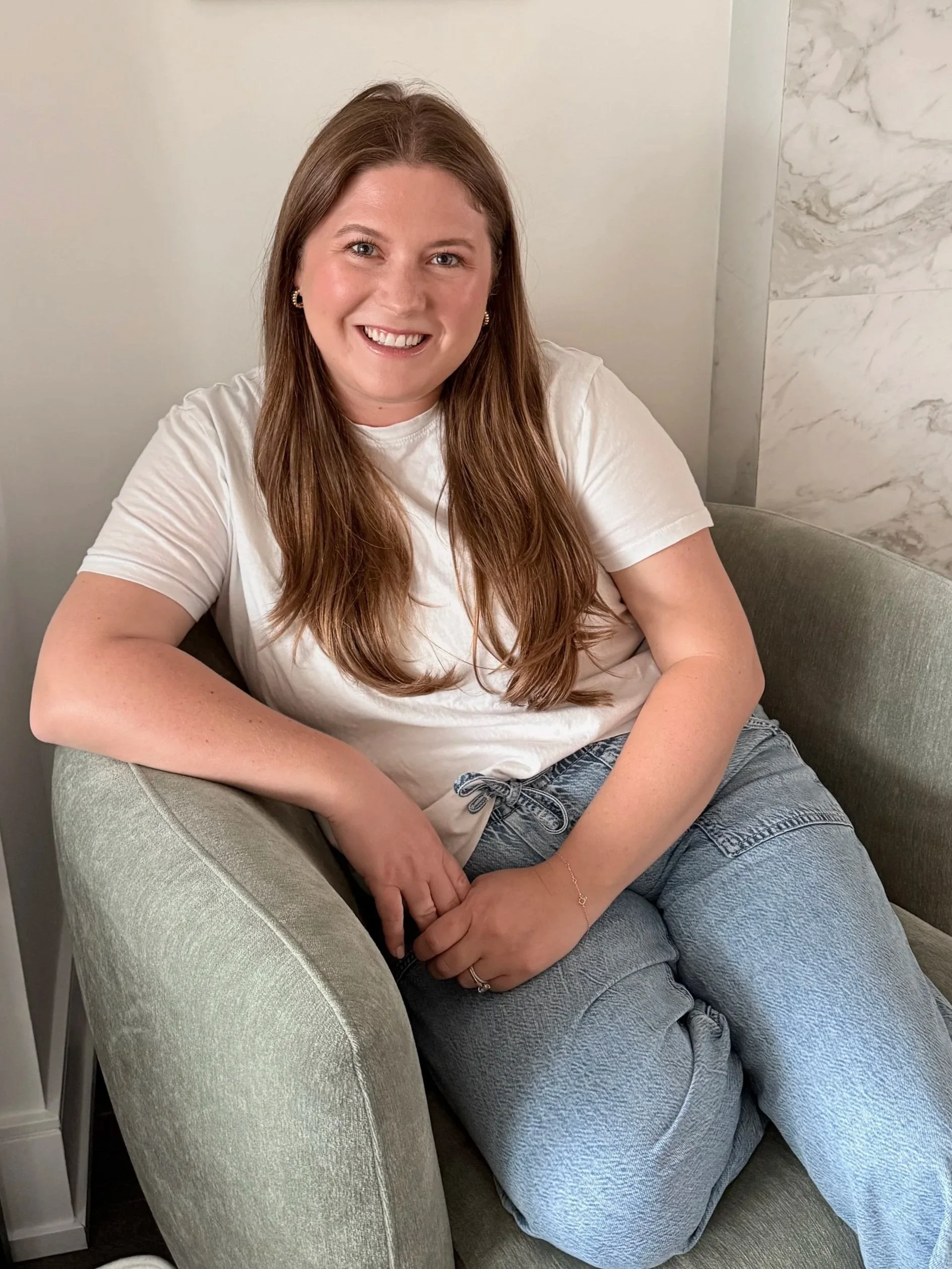 A woman with long brown hair, wearing a white t-shirt and gray sweatpants, sitting comfortably on a light green armchair in a room with marble walls and a white wall in the background, smiling at the camera.