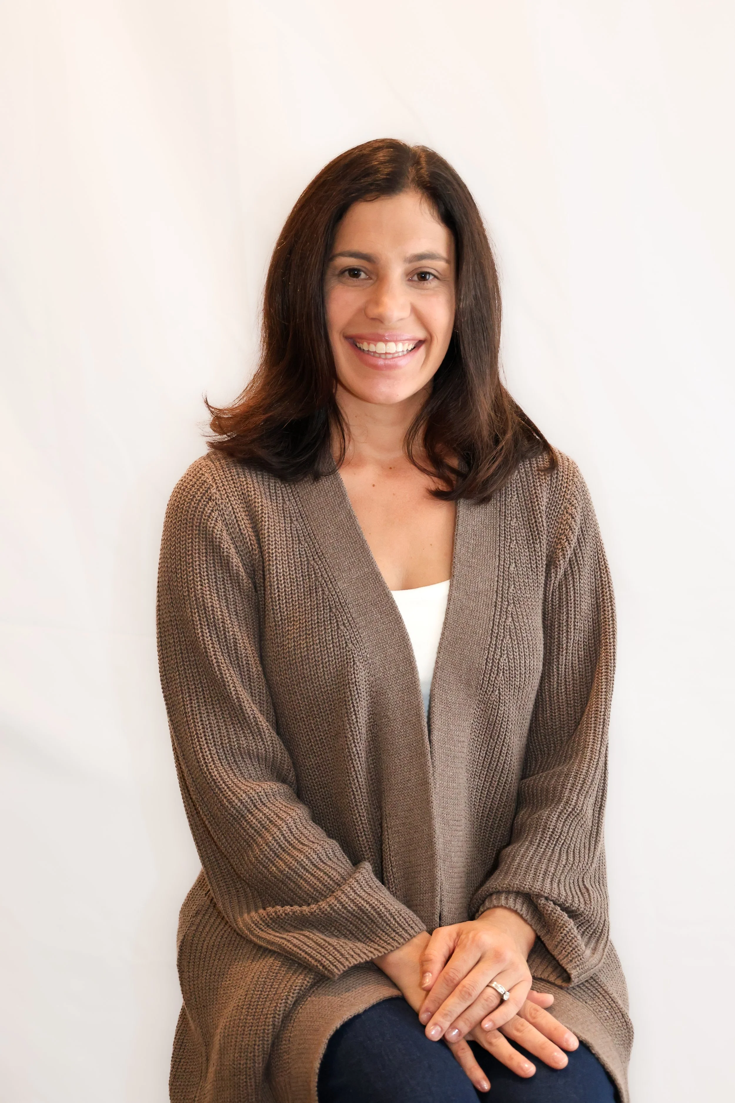 A woman with shoulder-length brown hair wearing a brown cardigan over a white top, sitting with her hands clasped on her lap, smiling, against a plain white background.