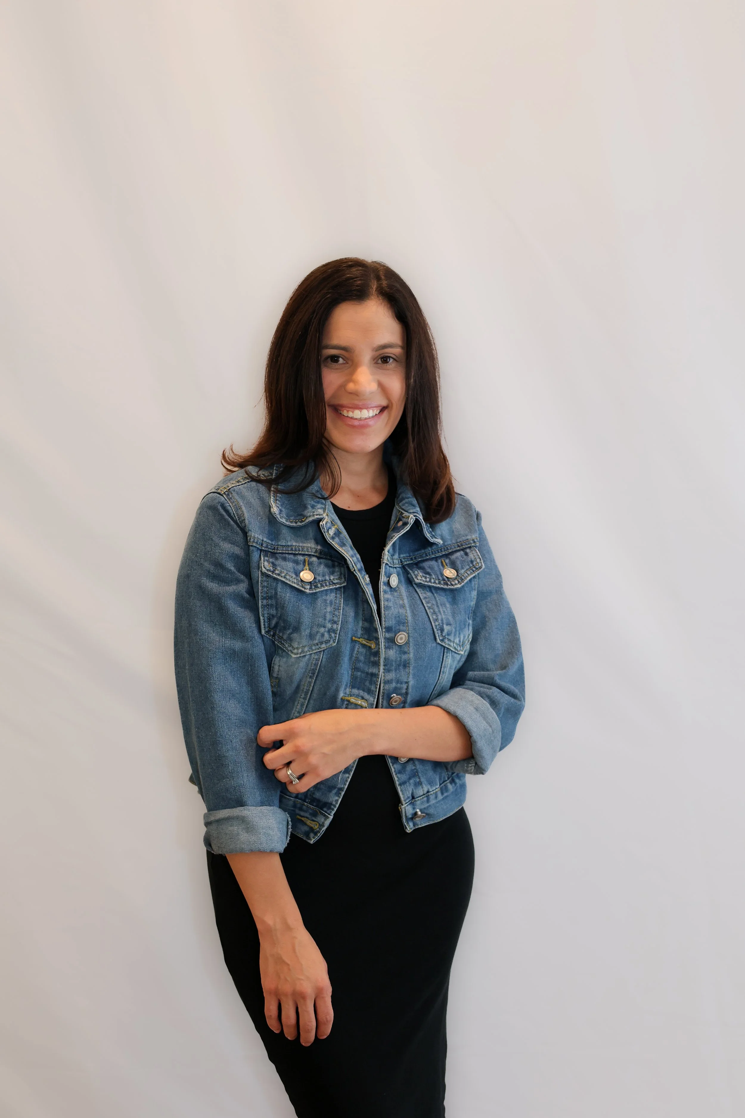 A woman with dark brown hair smiling, wearing a black dress and a denim jacket, standing against a plain white wall.
