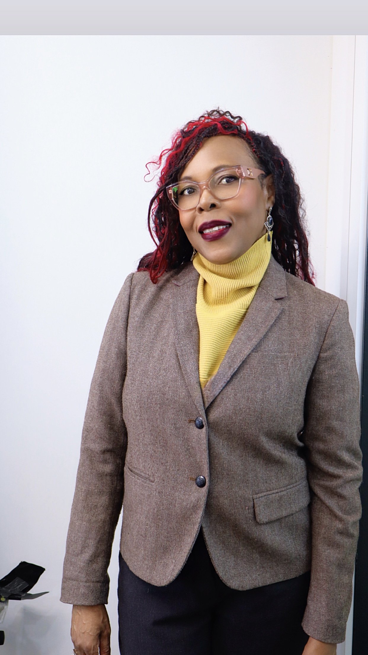 A woman with red and black curly hair, wearing glasses, earrings, a yellow turtleneck sweater, and a brown blazer, standing against a white background.