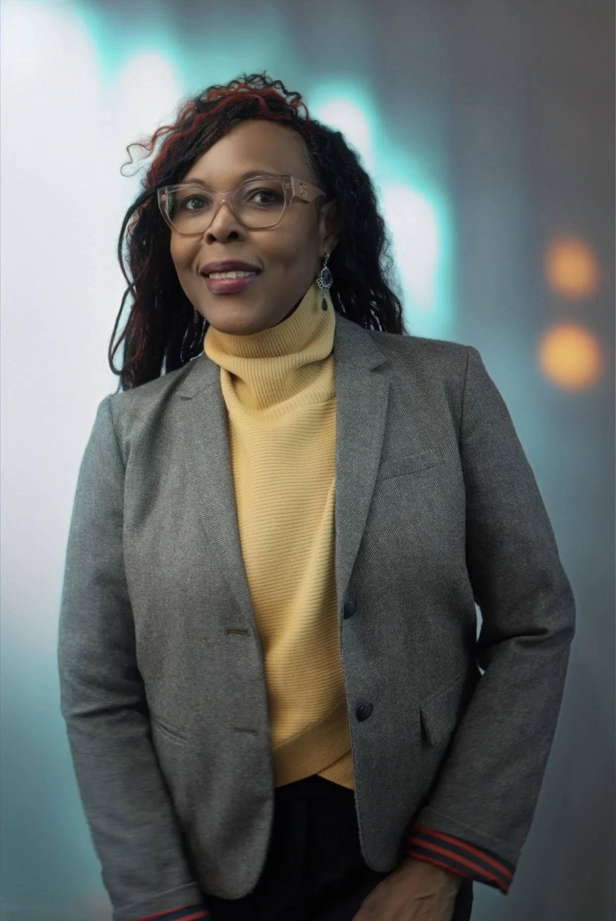 A woman with black and red curly hair, wearing glasses, earrings, a yellow turtleneck sweater, and a gray blazer, standing in front of blurred lights and wall background.