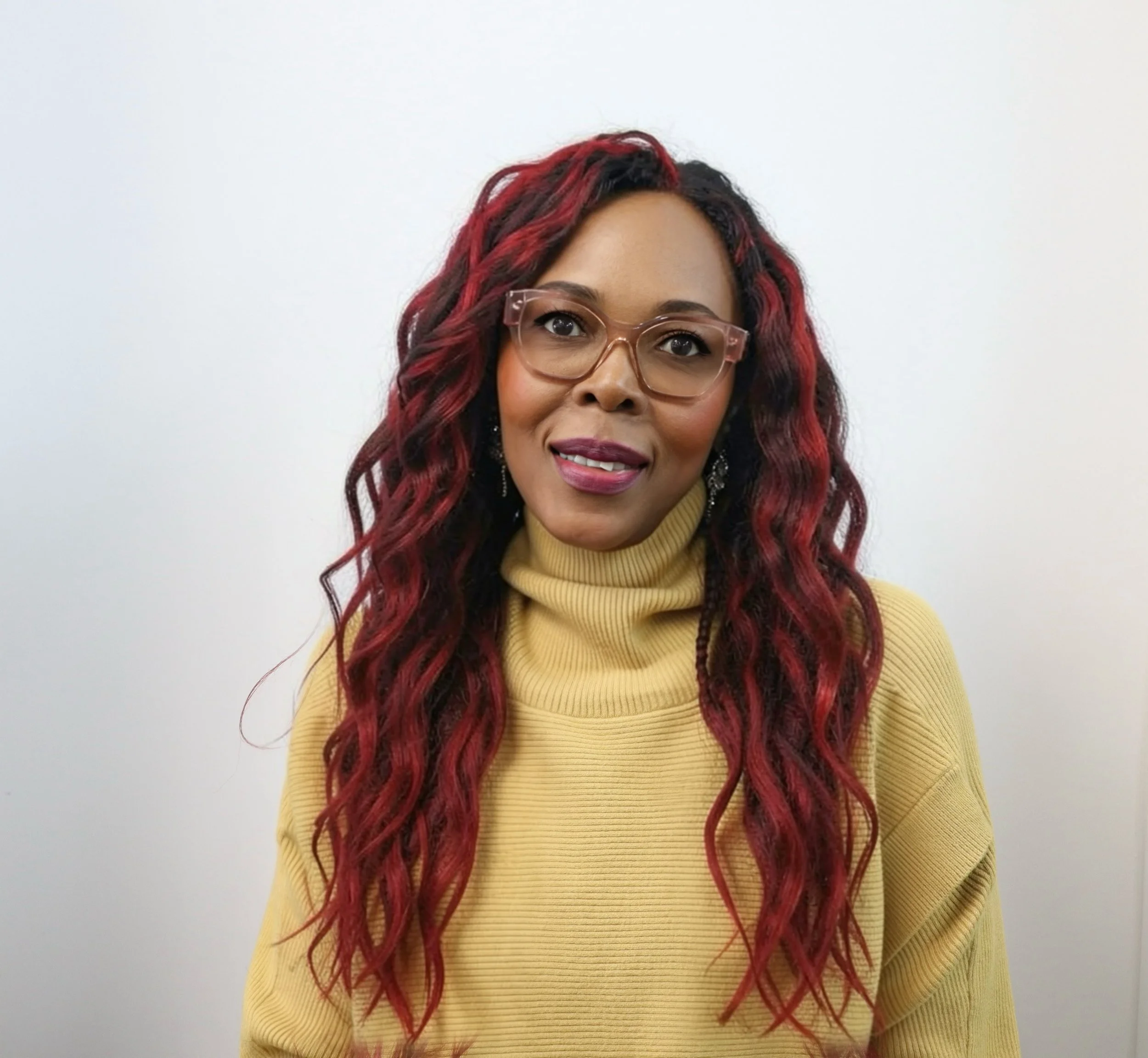 Close-up of a woman with curly red and black hair, wearing glasses and a mustard yellow turtleneck sweater, smiling in front of a plain white background.