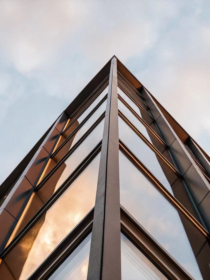 Photo d'un bâtiment moderne vu de bas, avec des fenêtres en verre et une structure en métal, reflétant le ciel.