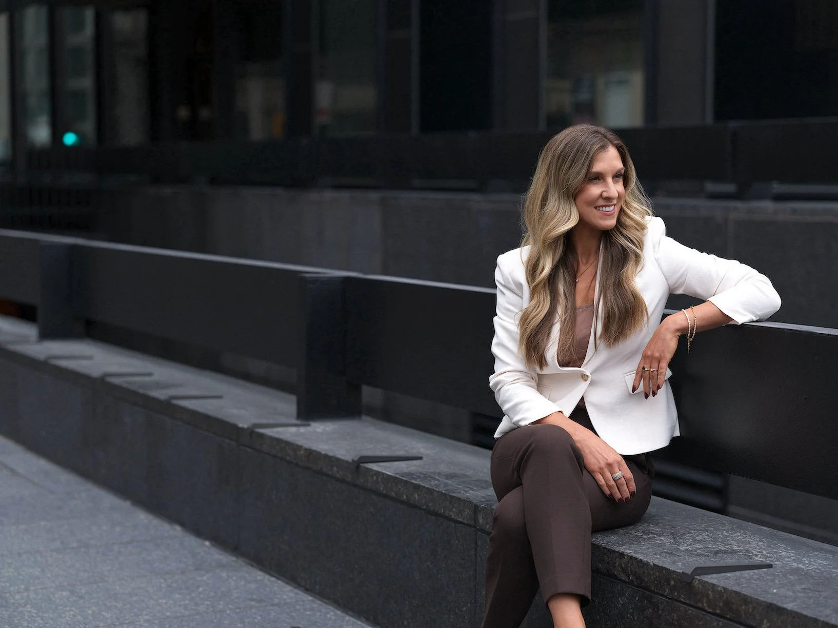 Une femme assise sur un banc noir en ville, portant une veste blanche, un pantalon marron, et souriant.