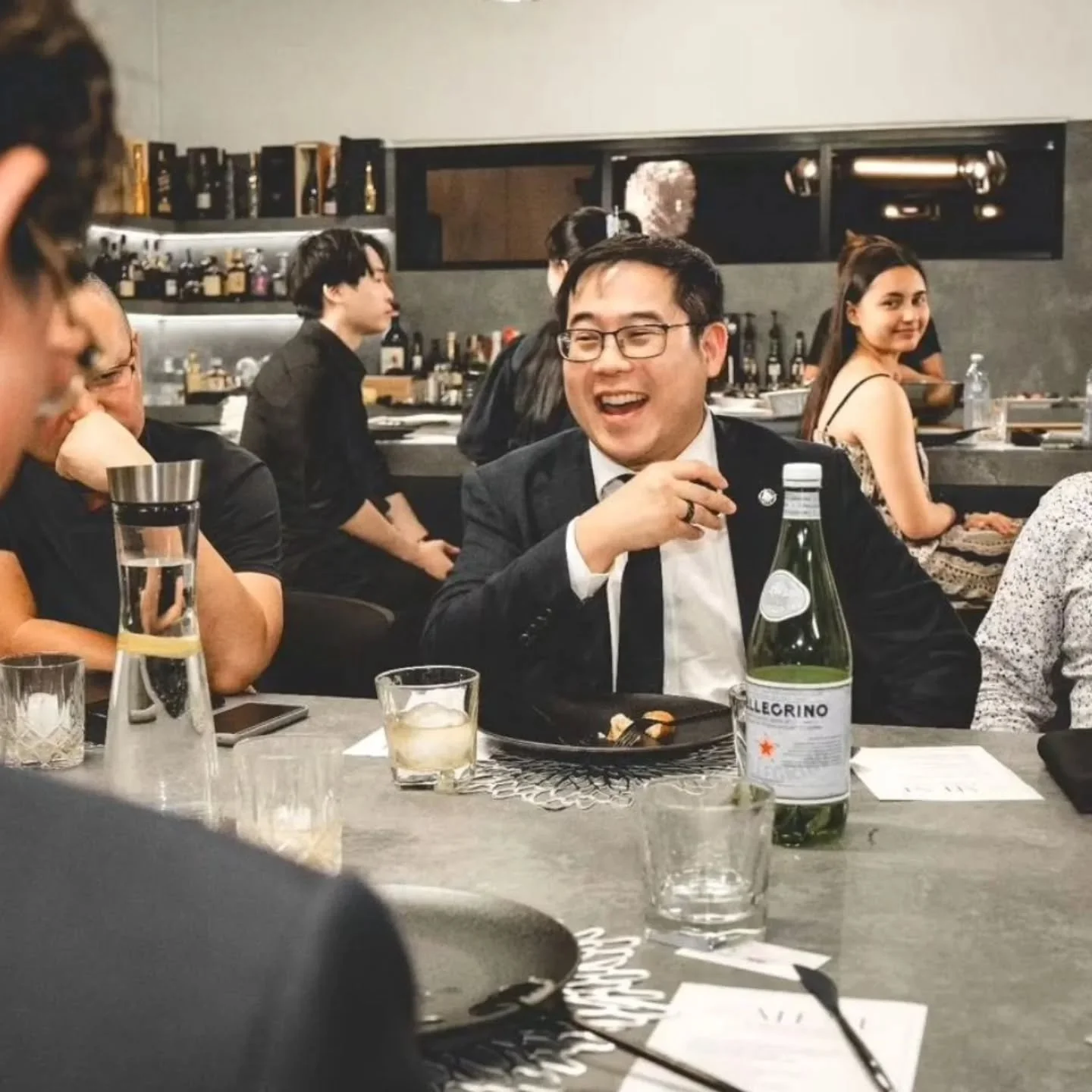 People seated at a restaurant table, smiling and engaging in conversation, with drinks including a large water bottle, glasses, and a plate of food on the table.