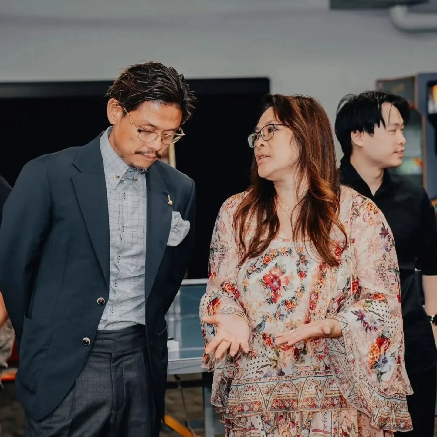 A man in a blue suit and glasses is looking down while a woman in a floral dress and glasses is speaking to him, gesturing with his hand. There is a young man in black in the background.