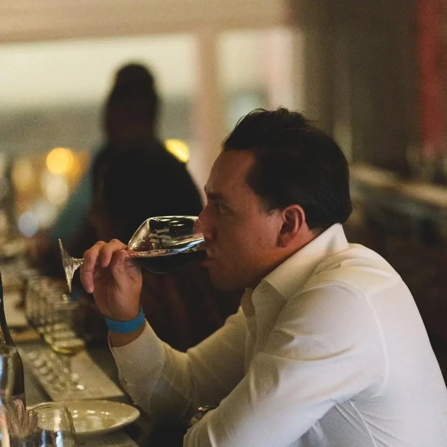 Man in a white shirt drinking red wine at a bar or restaurant, with blurred background and glasses on the table.