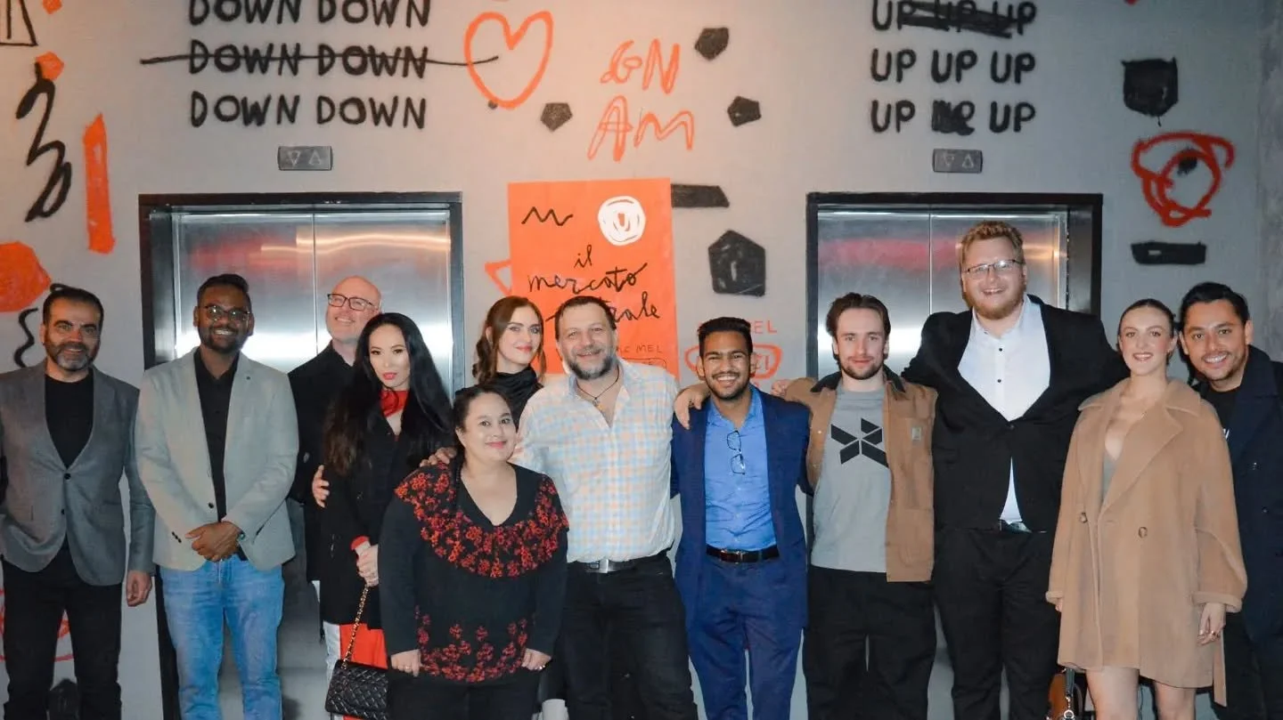 A group of fifteen people standing together in front of a wall with graffiti-style writing, including words like "DOWN" and "UP," and various drawings. The group is diverse, smiling, and dressed in a mix of casual and semi-formal attire.