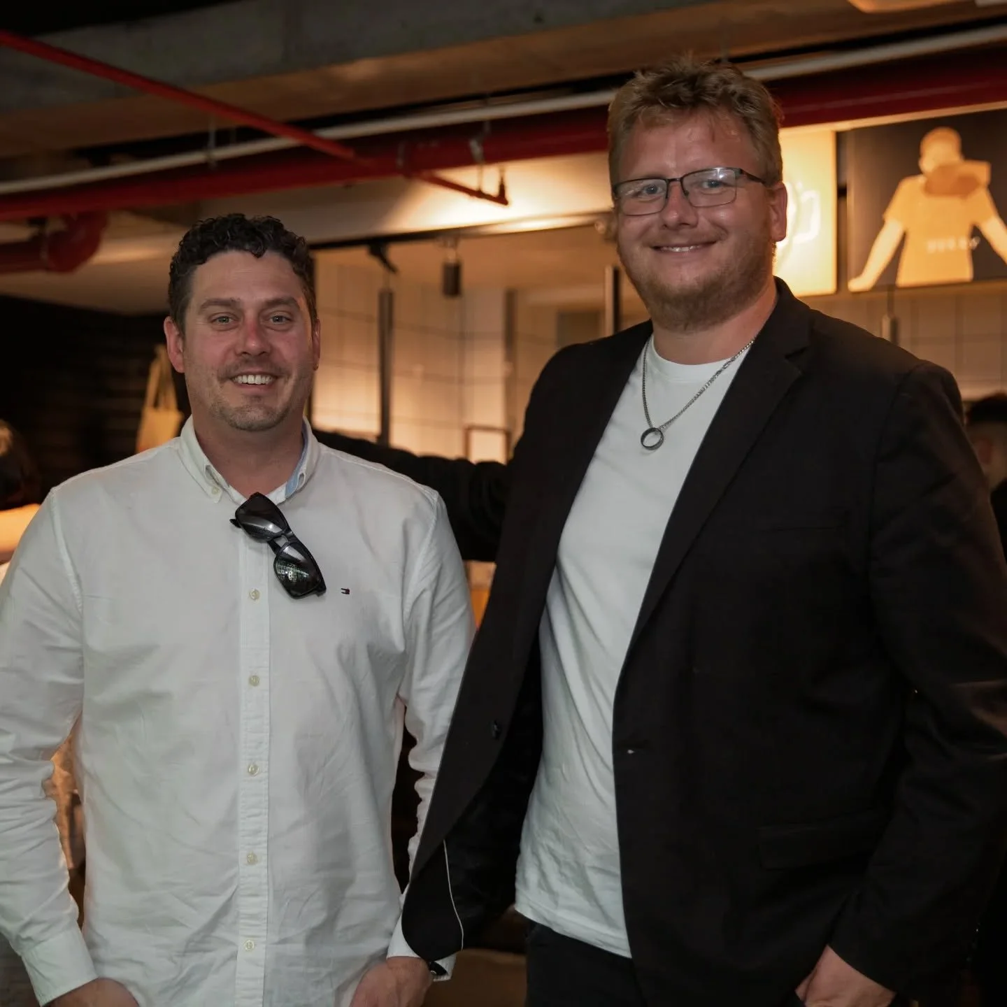 Two men smiling and standing close together in an indoor setting, with the man on the right wearing glasses, a black blazer, a white t-shirt, and a silver necklace, and the man on the left wearing a white button-up shirt with sunglasses hanging from the collar.