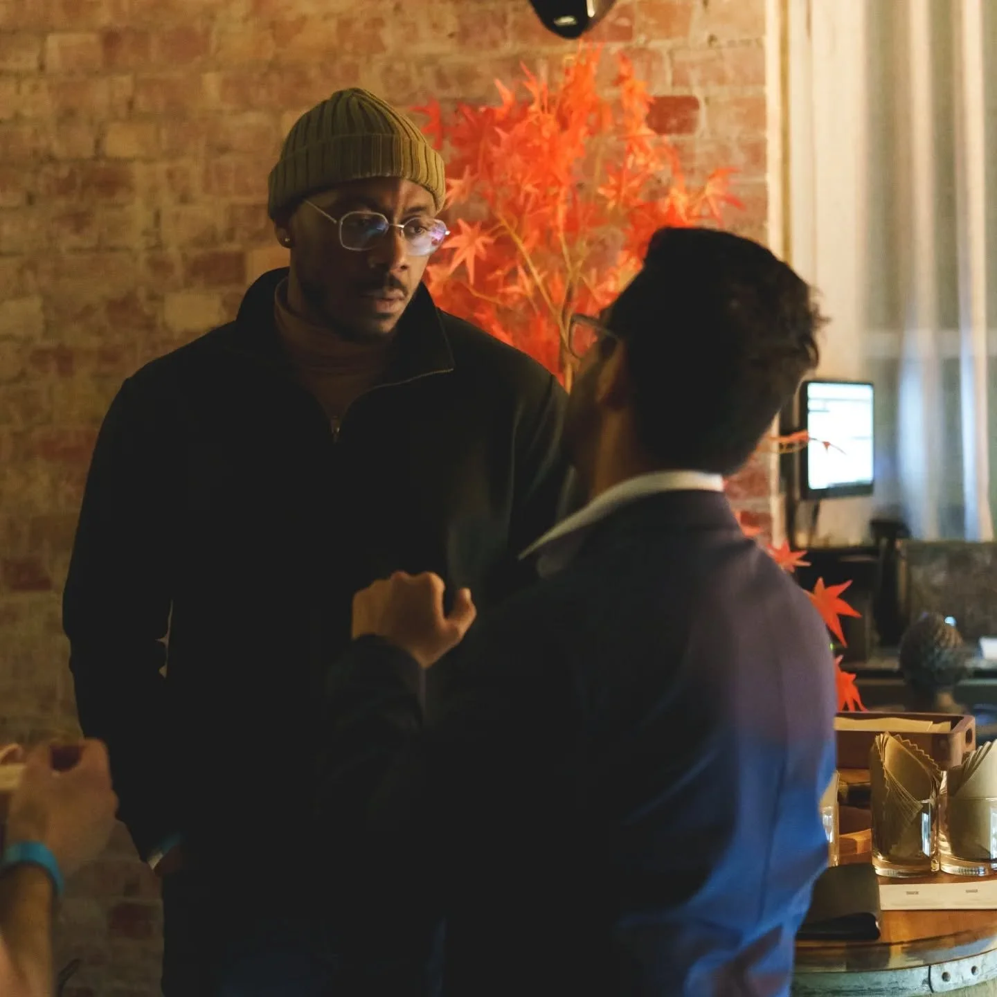 Two men with dark hair and glasses engaged in a conversation inside a cozy, dimly lit cafe with rustic brick walls and warm decor, including an orange autumn leaf tapestry and a computer on a desk in the background.
