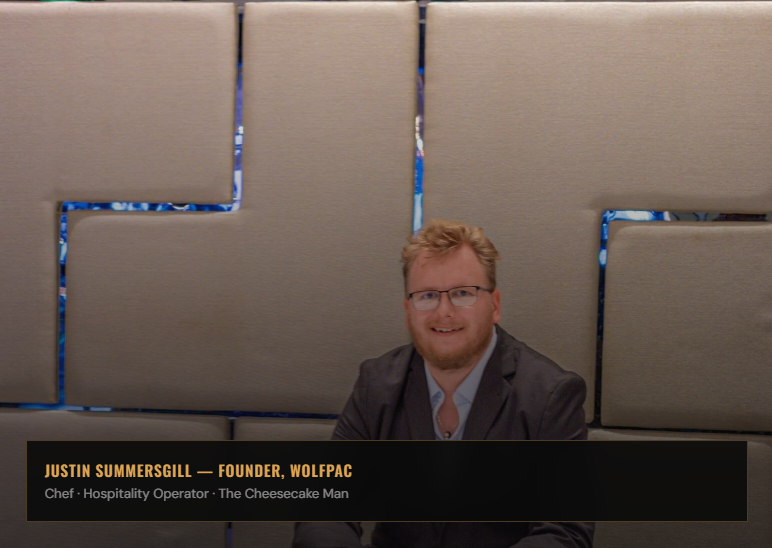 A man with glasses and a beard sitting in front of a beige wall with geometric cutouts, smiling at the camera.