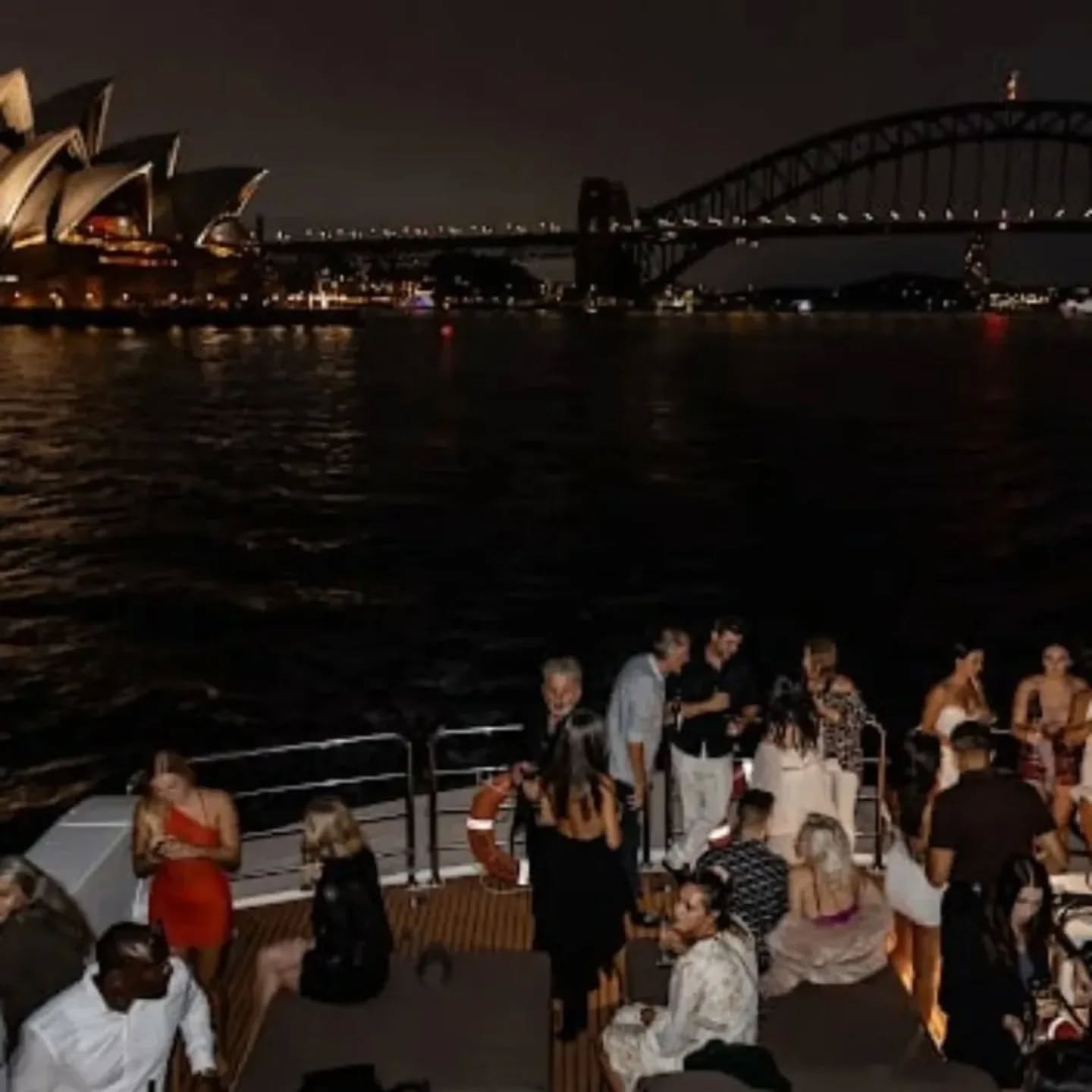 Nighttime scene on a boat with people gathered on the deck, a view of the Sydney Opera House and Harbour Bridge illuminated in the background.