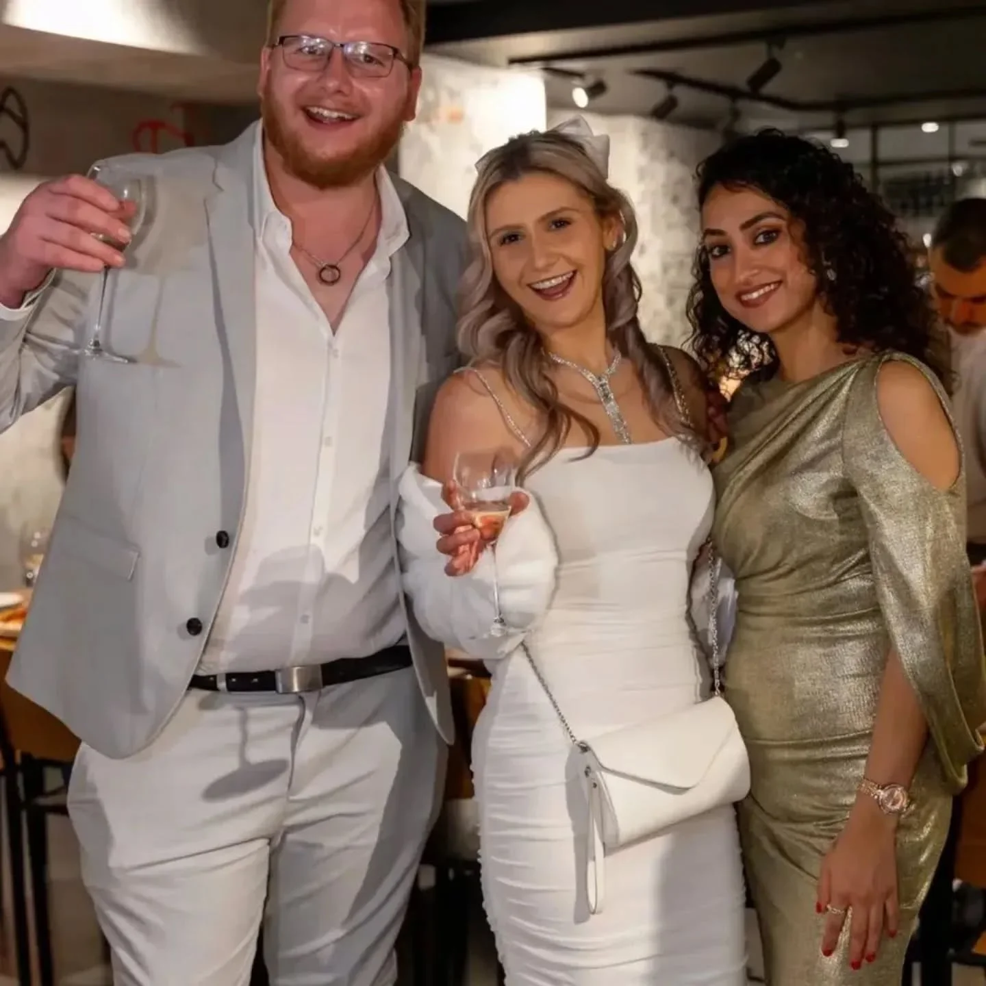 Group of three friends dressed in formal party attire, smiling, holding drinks, at a social gathering in a restaurant or bar.