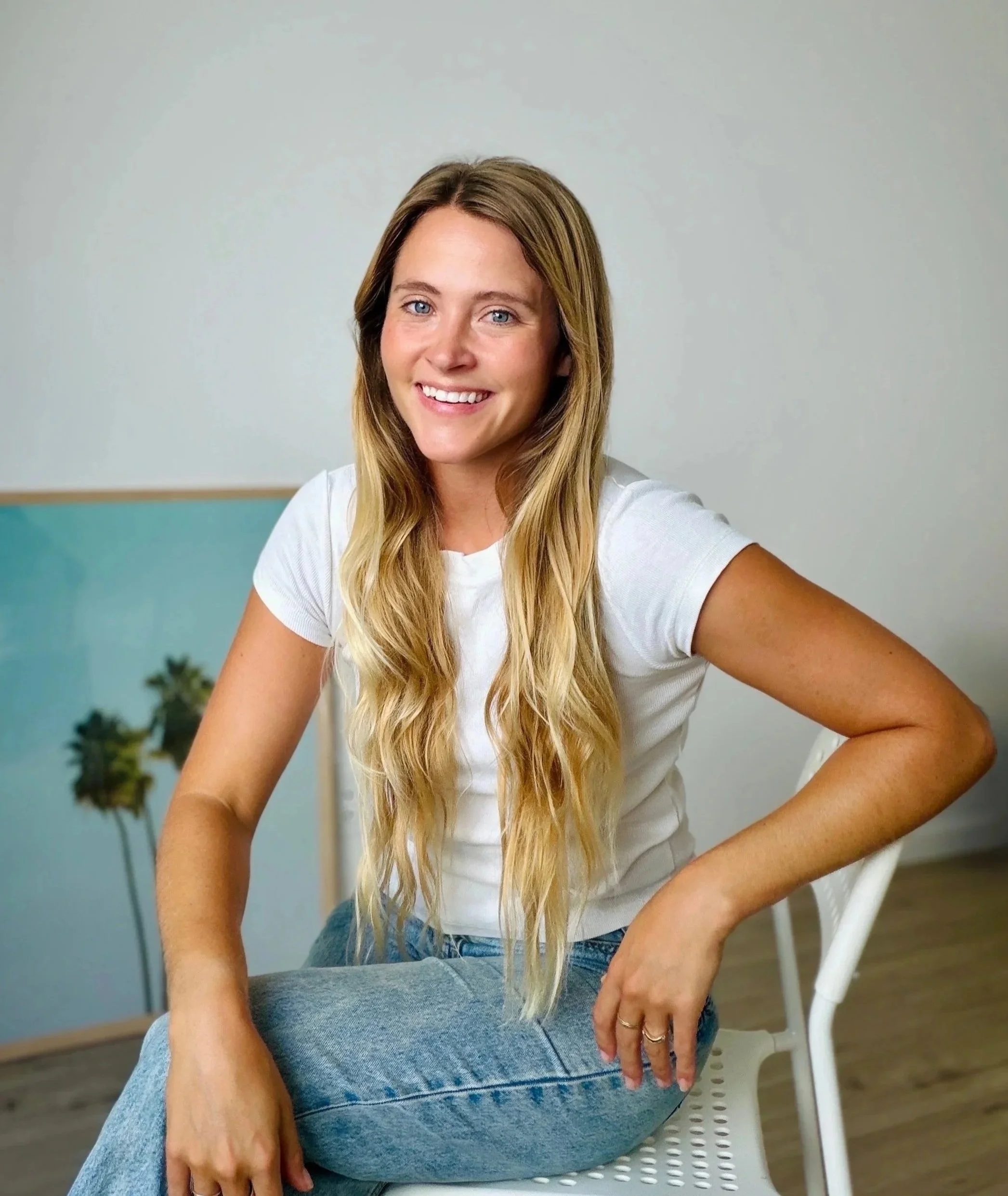 A young woman with long blonde hair, blue eyes, wearing a white t-shirt, sitting on a white chair, smiling at the camera, indoors with a plain light gray wall background and some outdoor trees visible at her occupational therapy clinic.