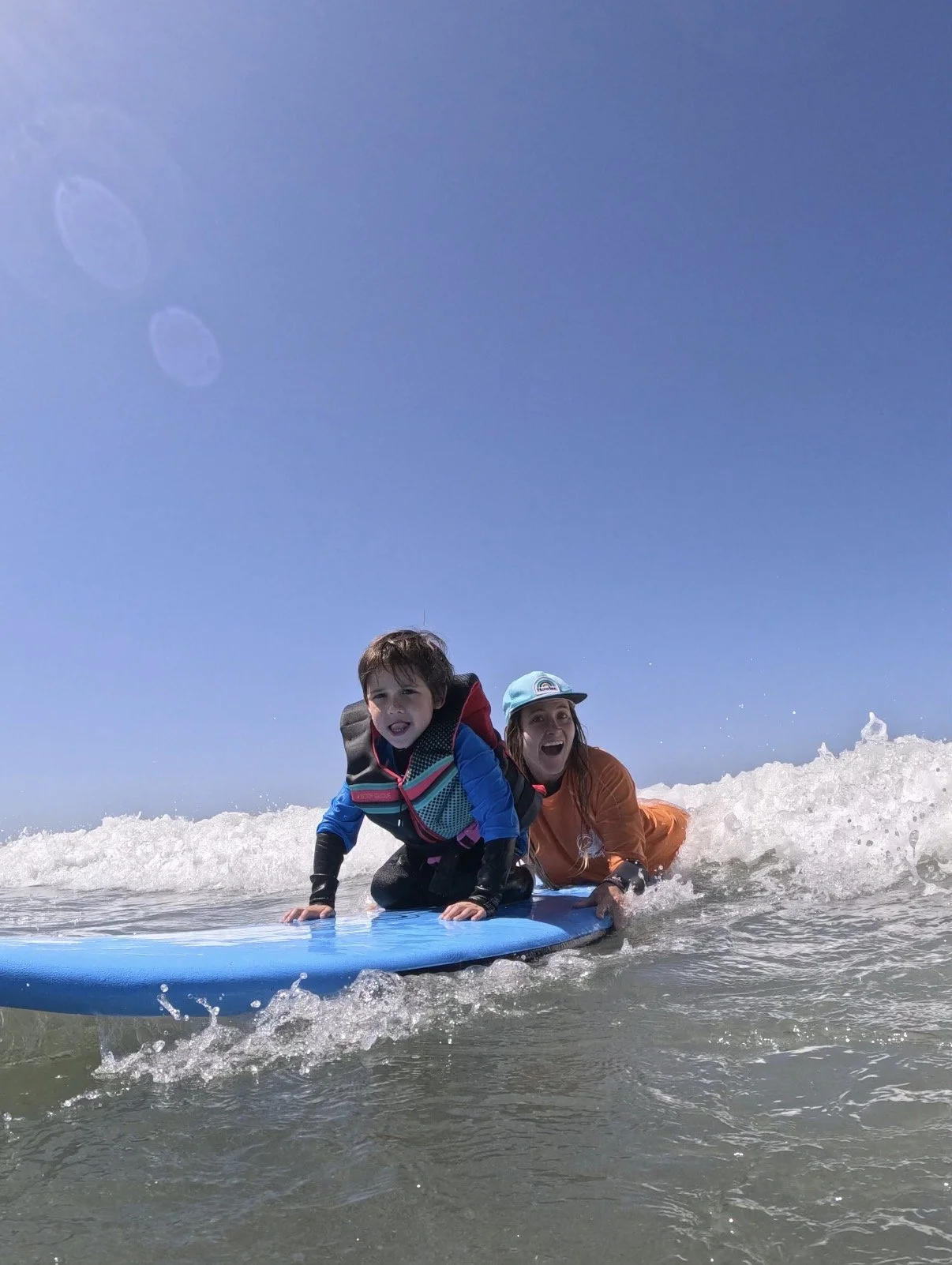 A woman and a young boy surfing on a blue surfboard in the ocean, with the woman holding onto the surfboard and both smiling as they enjoy the water under a clear blue sky during surf therapy with an occupational therapist. 