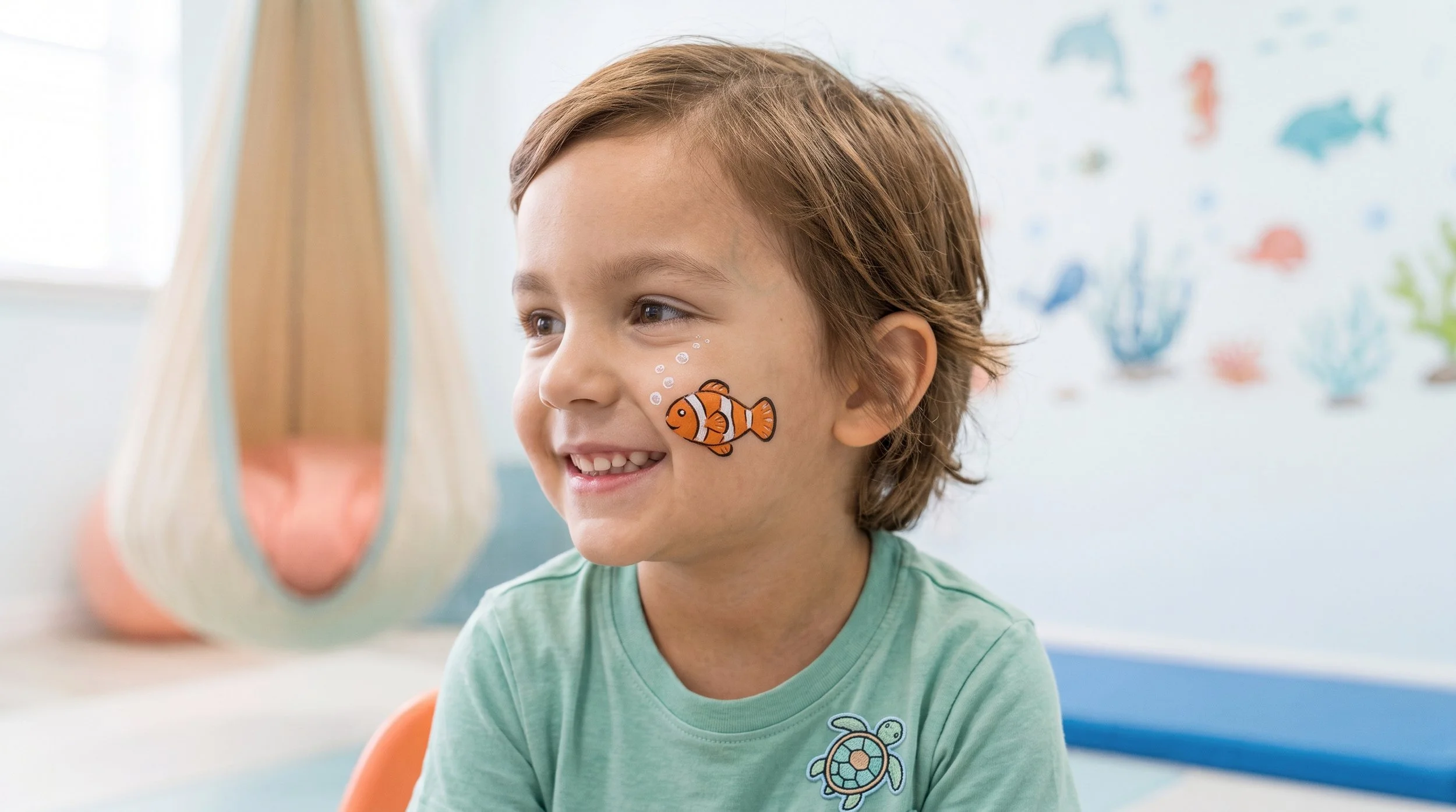 A young boy with light brown hair and a sticker of a clownfish on his cheek, smiling as he looks at an object outside the frame, in a room decorated with aquatic-themed wall art.
