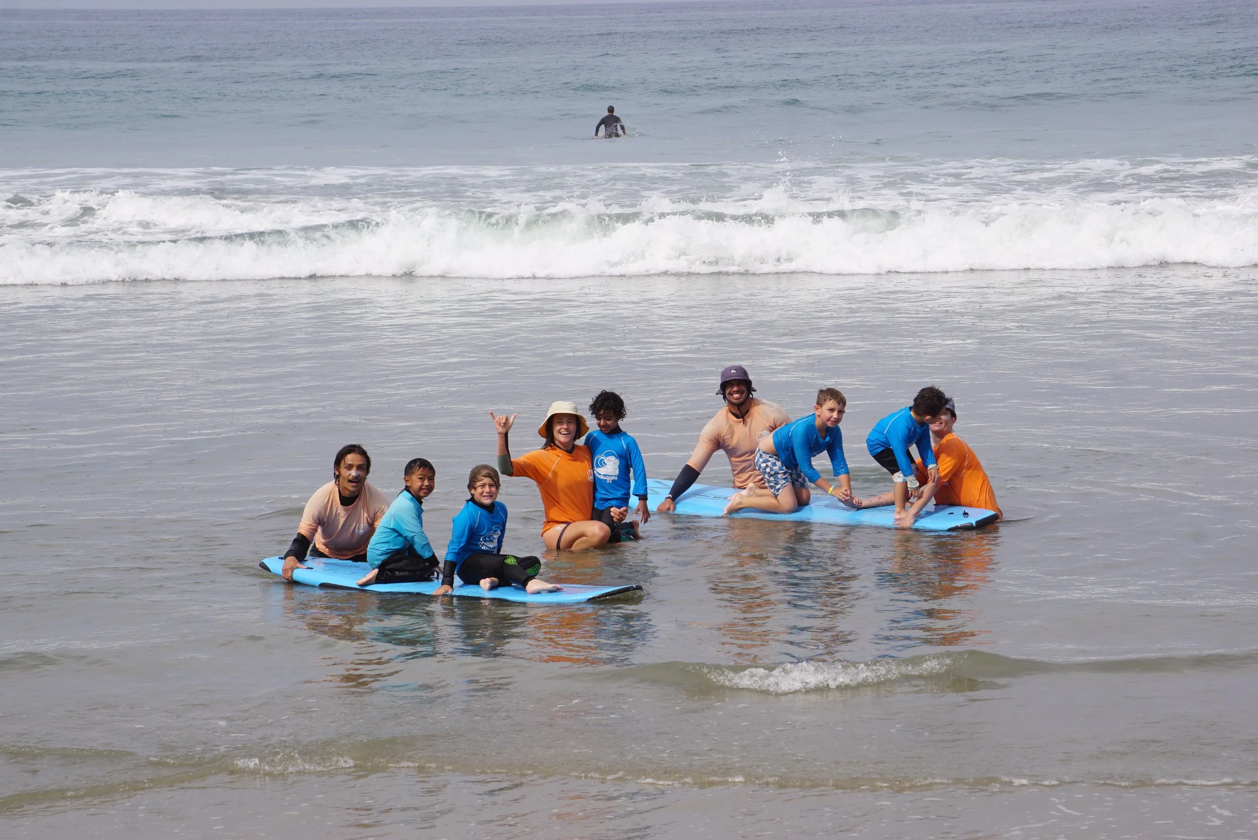 Group of children and adults on a surfboard in shallow ocean water, smiling and posing for a photo with waves and a person surfing in the background during surf therapy with an occupational therapist.