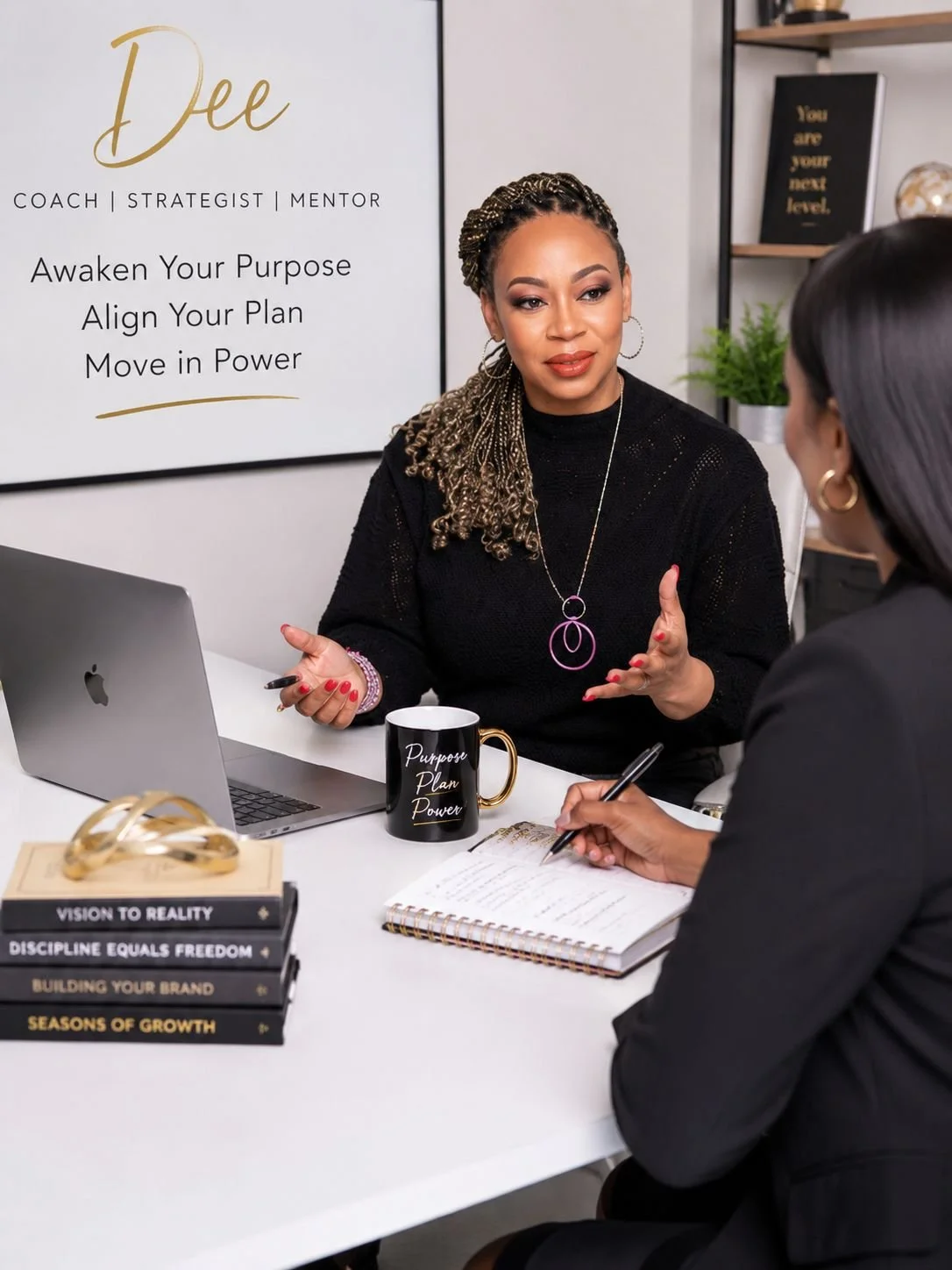 Two women having a coaching session at a desk, with a sign behind them reading "Dee Coach, Strategist, Mentor," and a stack of books on personal development.